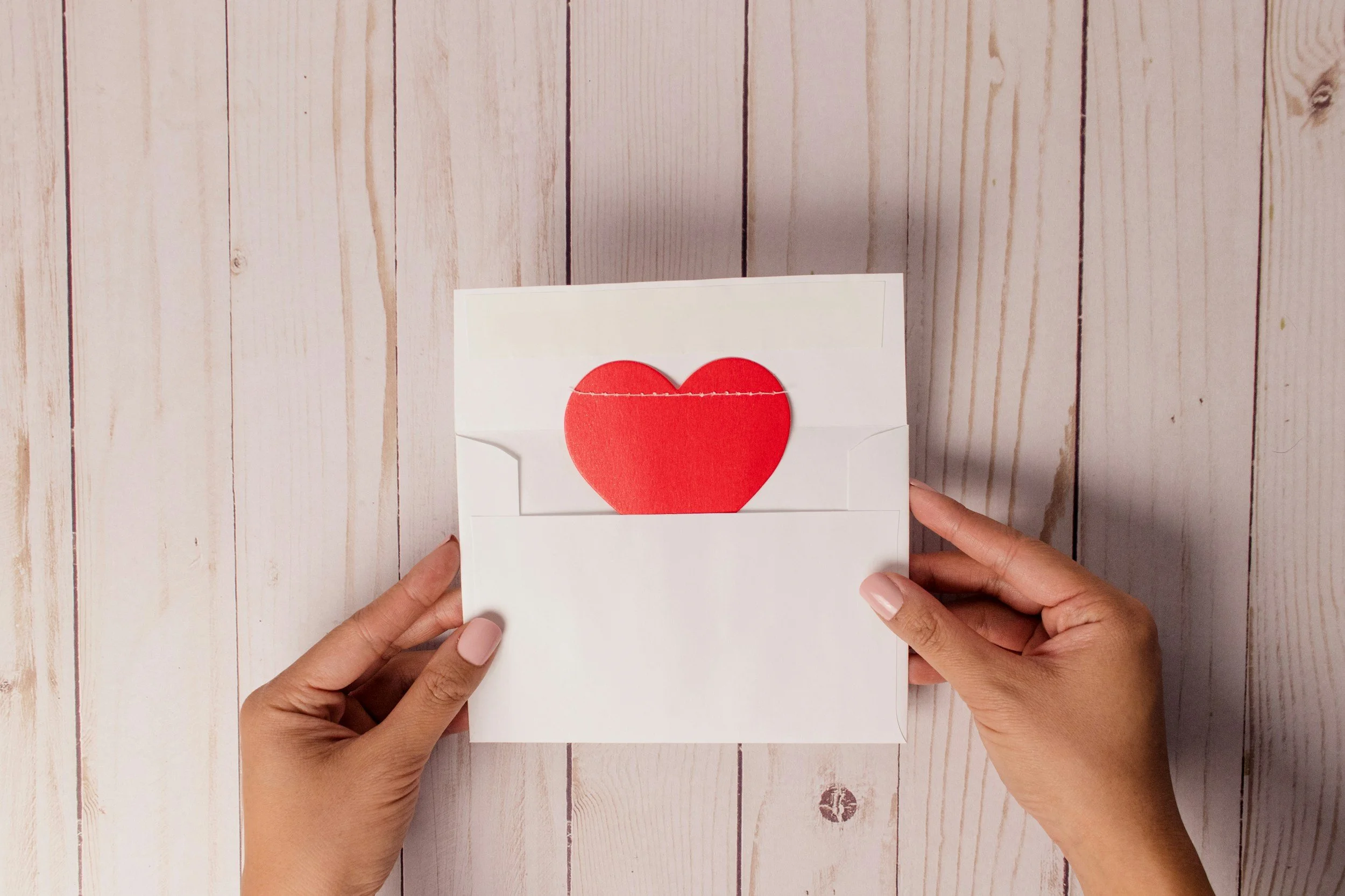 Person holding an open envelope with a red heart inside on a light wooden surface.