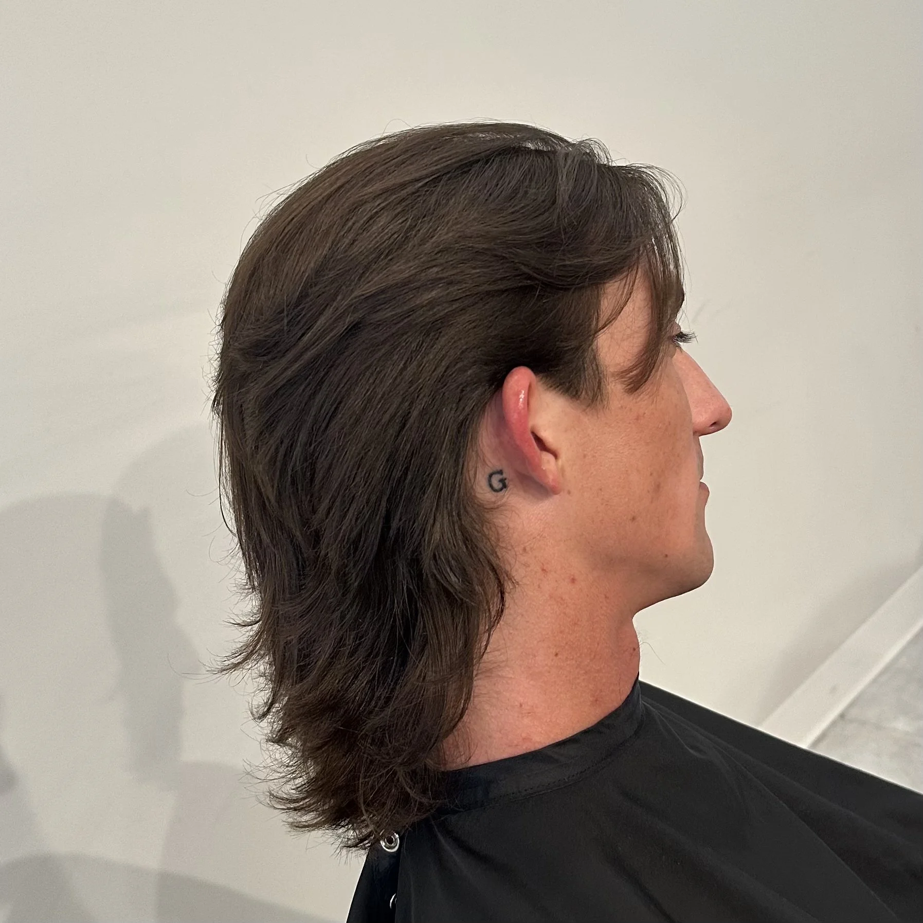 Profile view of a man with long brown hair and a small tattoo of the letter G behind his ear, sitting in a salon or barbershop.