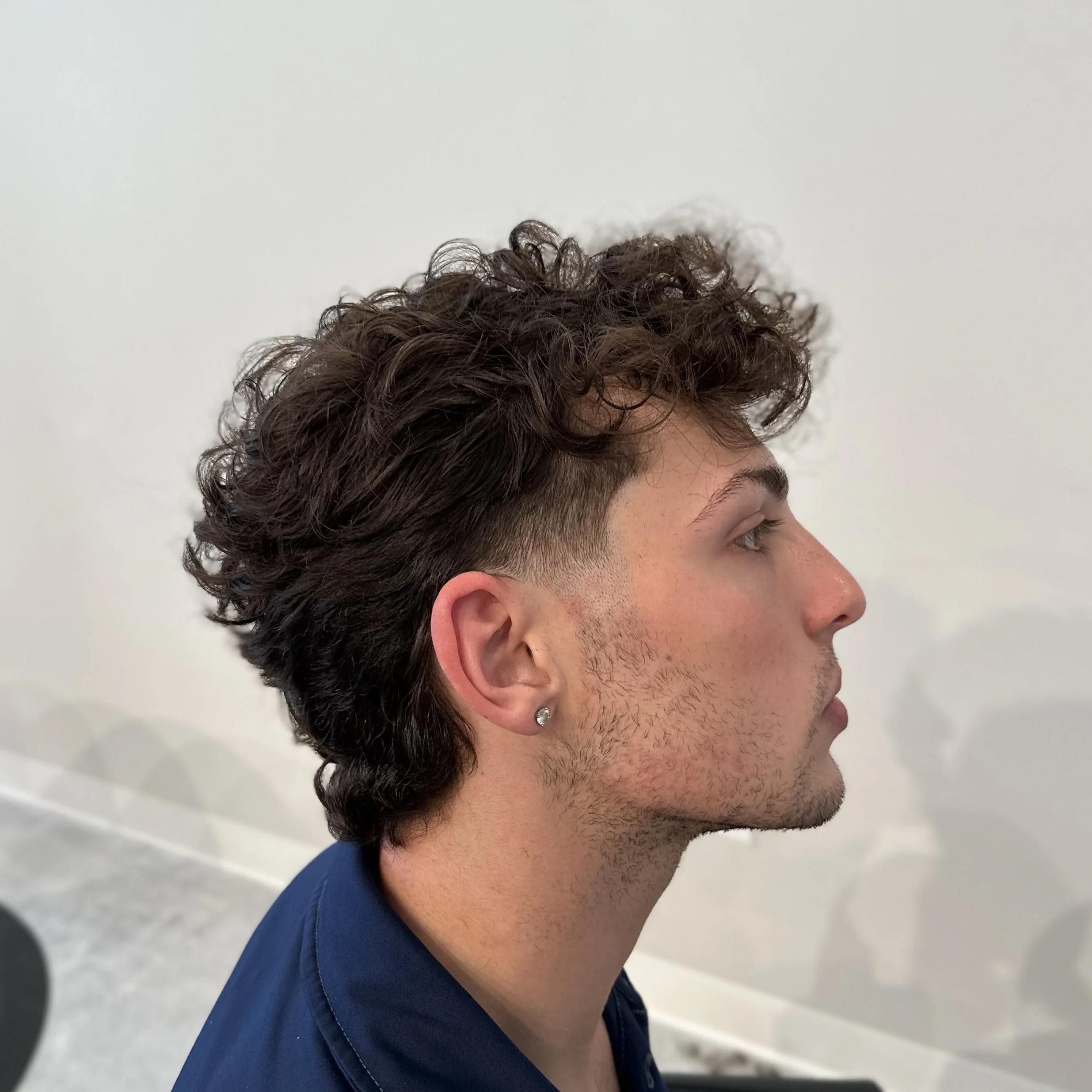 Profile of a young man with curly brown hair, wearing a blue shirt and a small earring, against a white background.
