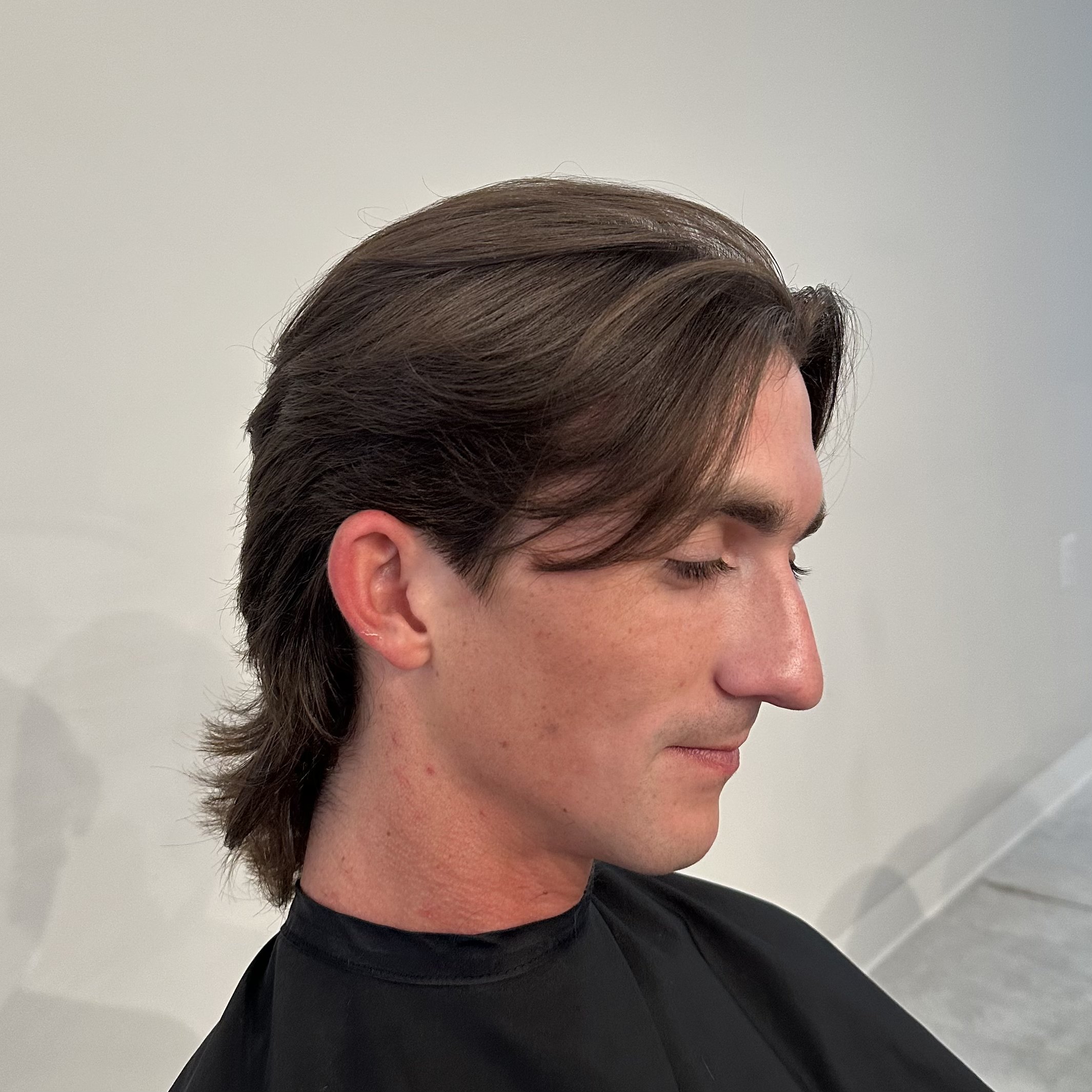 Profile of a young man with brown hair, closed eyes, and light skin, wearing a black cape, against a plain light-colored background.