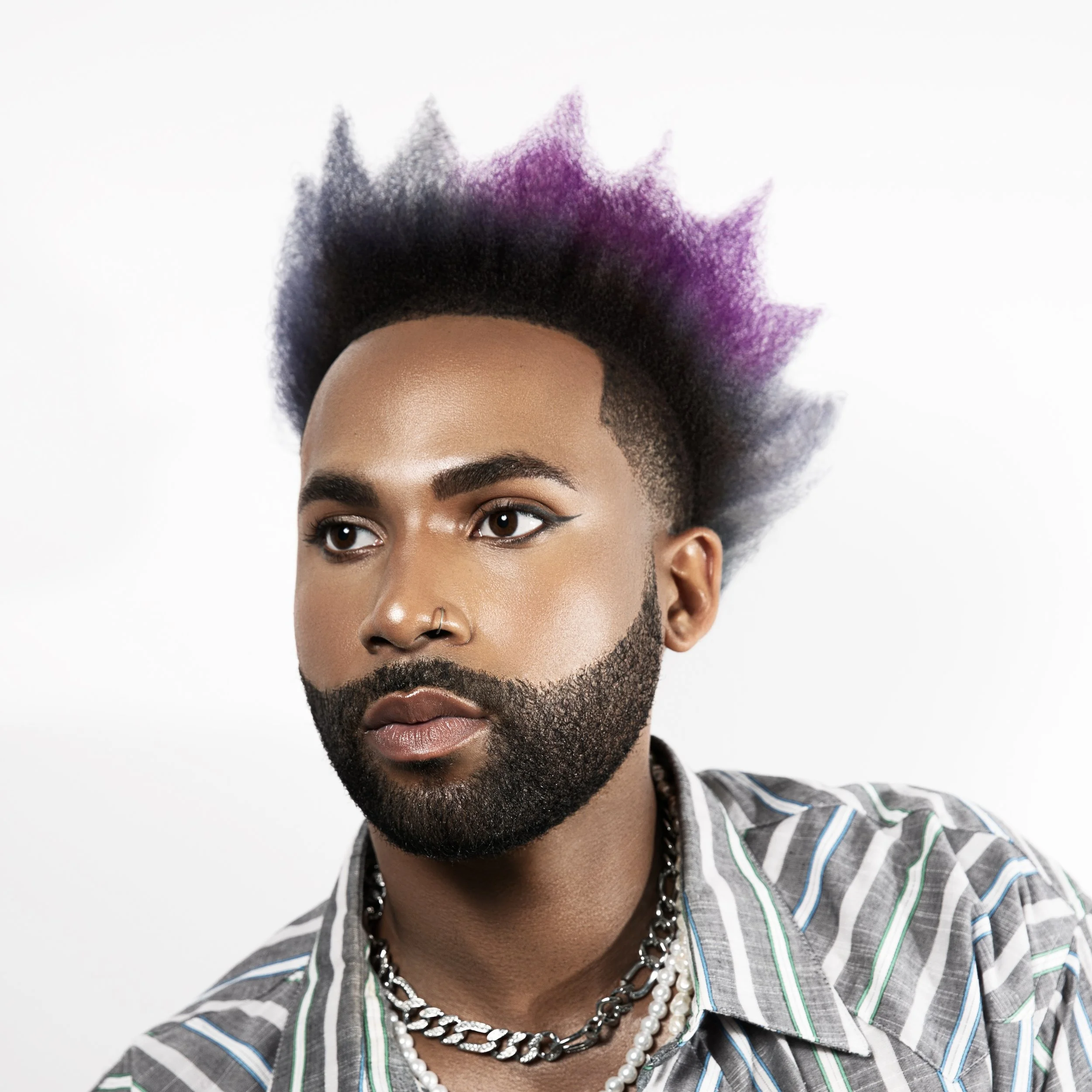 A person with a vibrant mohawk hairstyle featuring black and purple colors, groomed facial hair, eyeliner, and accessorized with layered chains and necklaces, wearing a striped shirt.