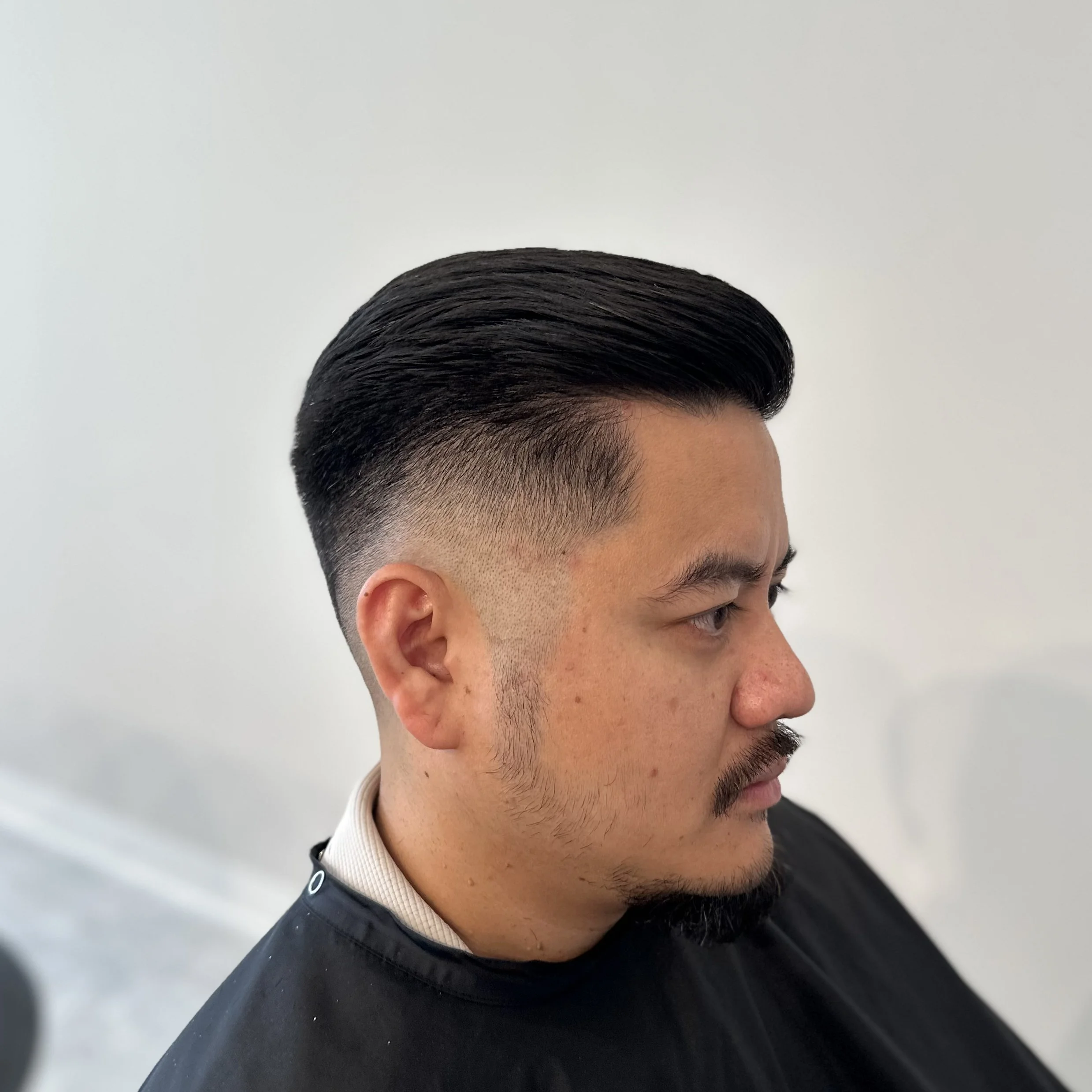 Side view of a man with a modern fade haircut, dark hair styled back, and a trimmed beard, sitting in a salon.