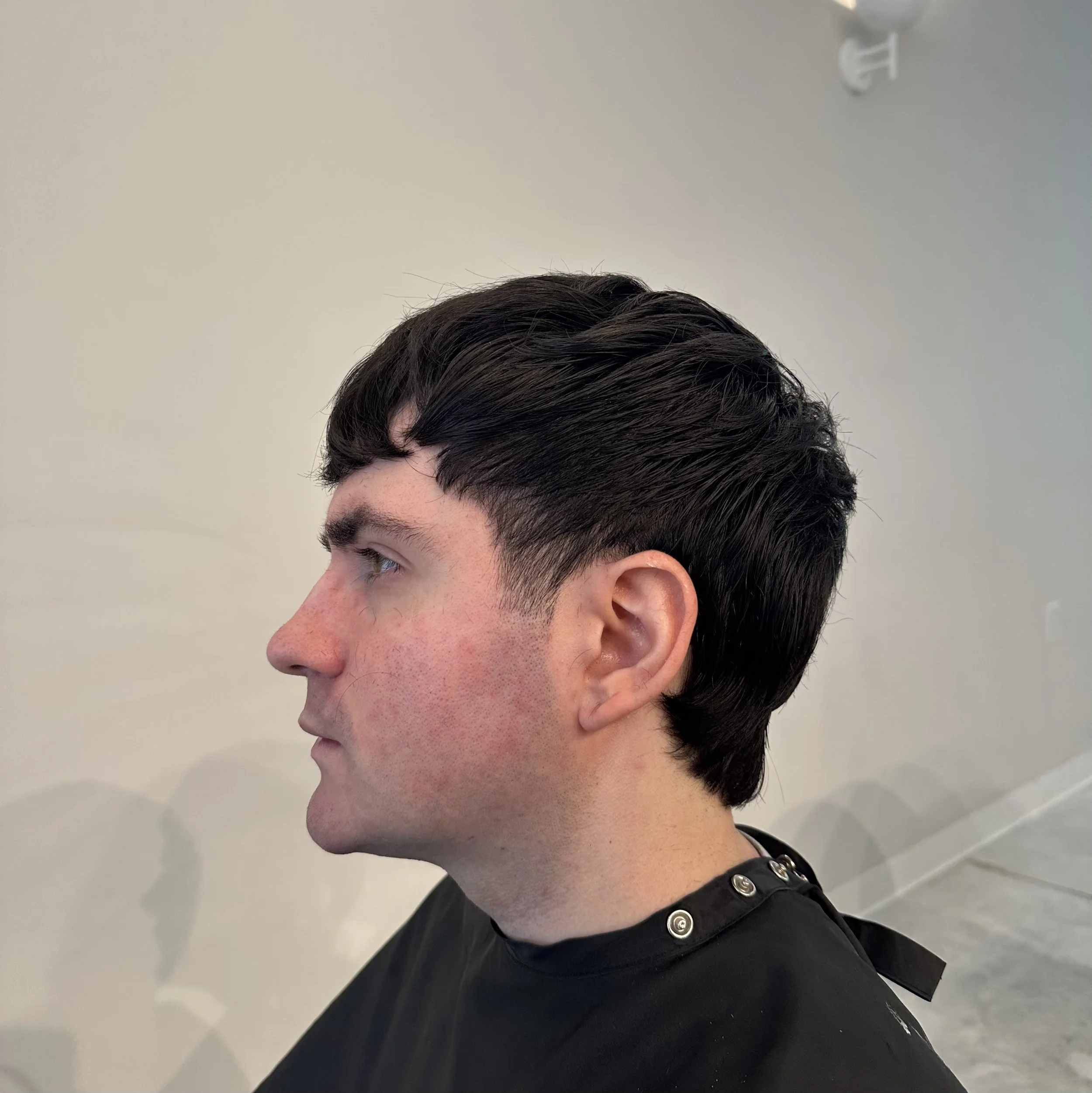 Profile of a young man with dark, short, styled hair wearing a black barber cape in a salon or barbershop.