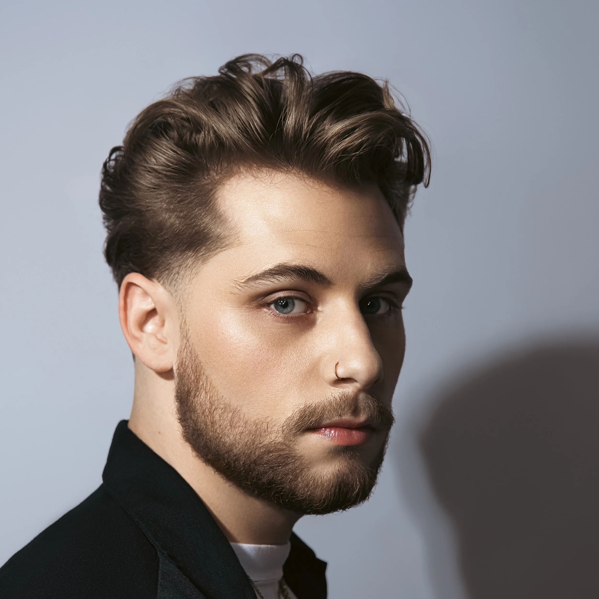 Close-up portrait of a young man with wavy brown hair, blue eyes, and a well-groomed beard, wearing a black jacket against a plain background.