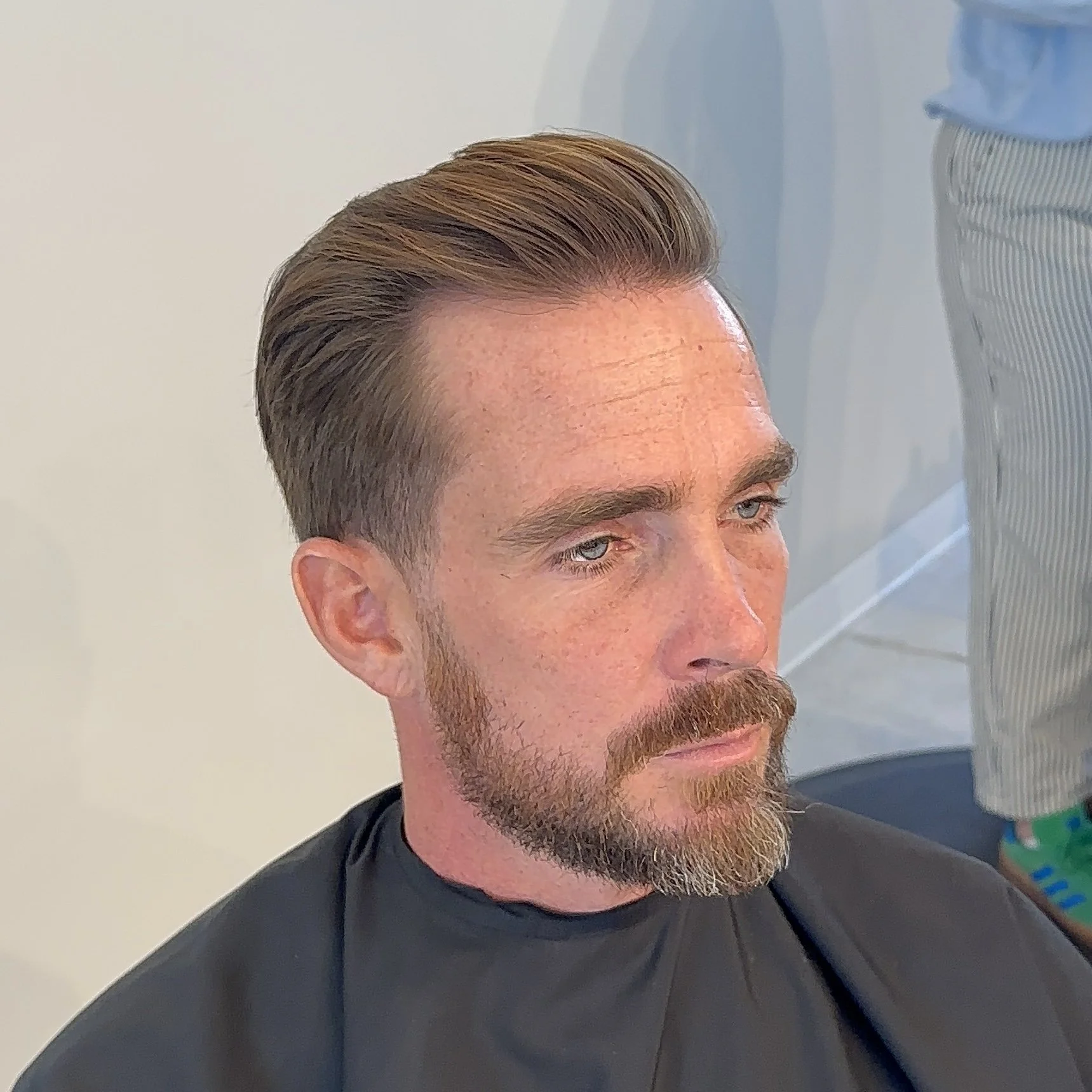 Man with neatly styled brown hair, a beard, and a mustache, sitting in a barbershop with a black cape around his shoulders.