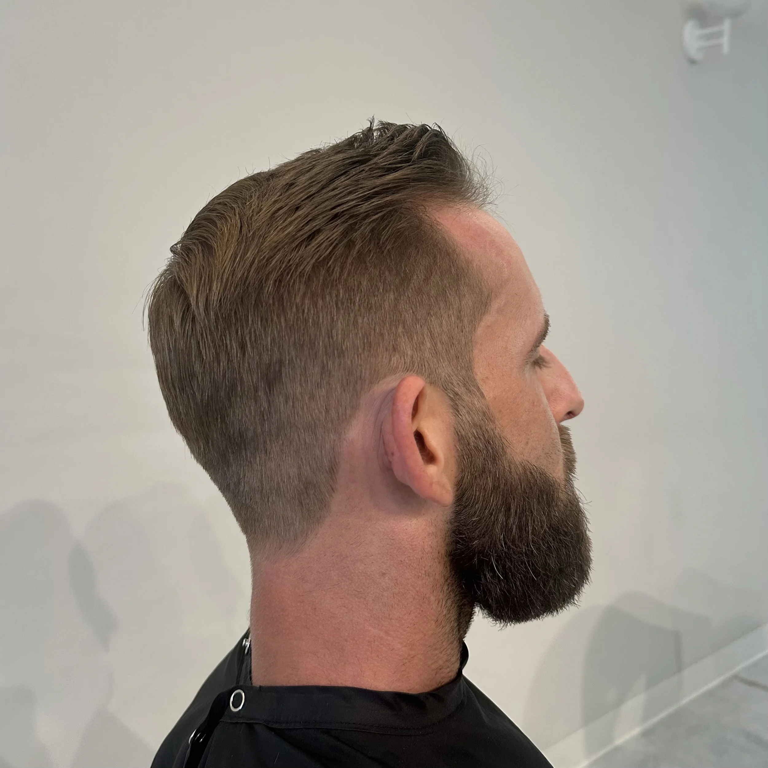 Side view of a man with a fresh haircut and beard, sitting in a salon chair against a plain wall.