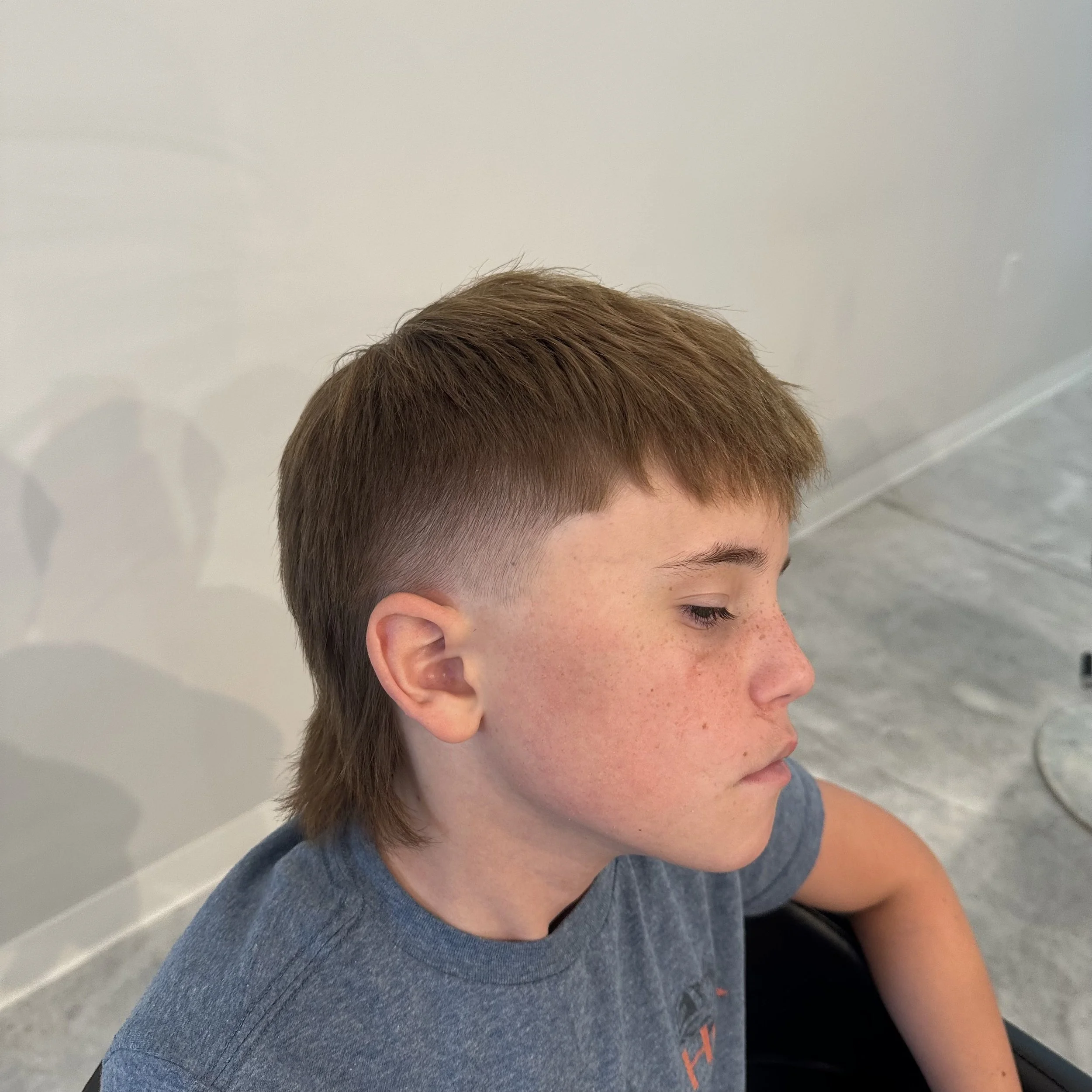 Side profile of a young boy with medium-length brown hair sitting indoors on a gray floor and plain light-colored wall