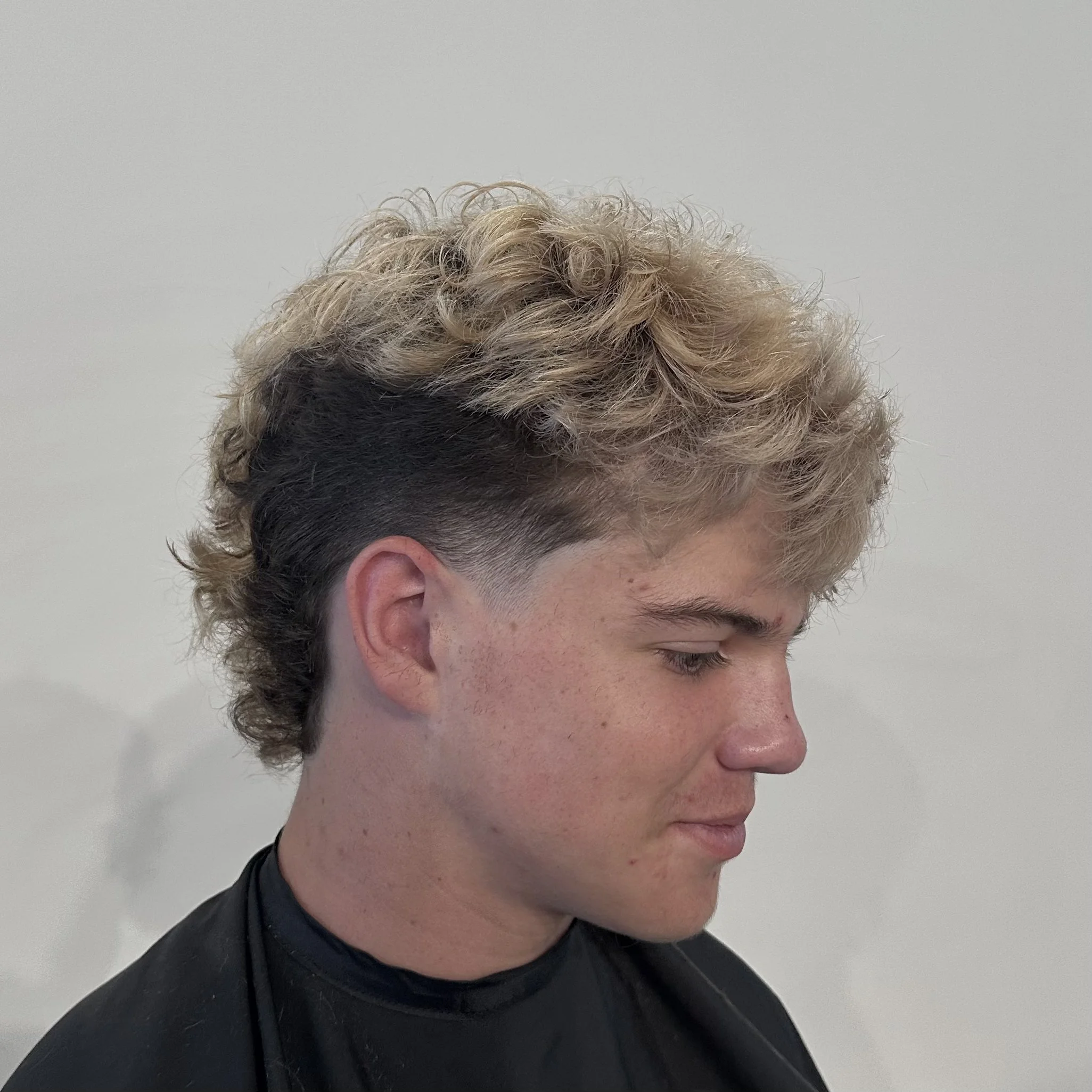 Young man with a curly, blonde and black undercut hairstyle, looking down, wearing a black shirt.