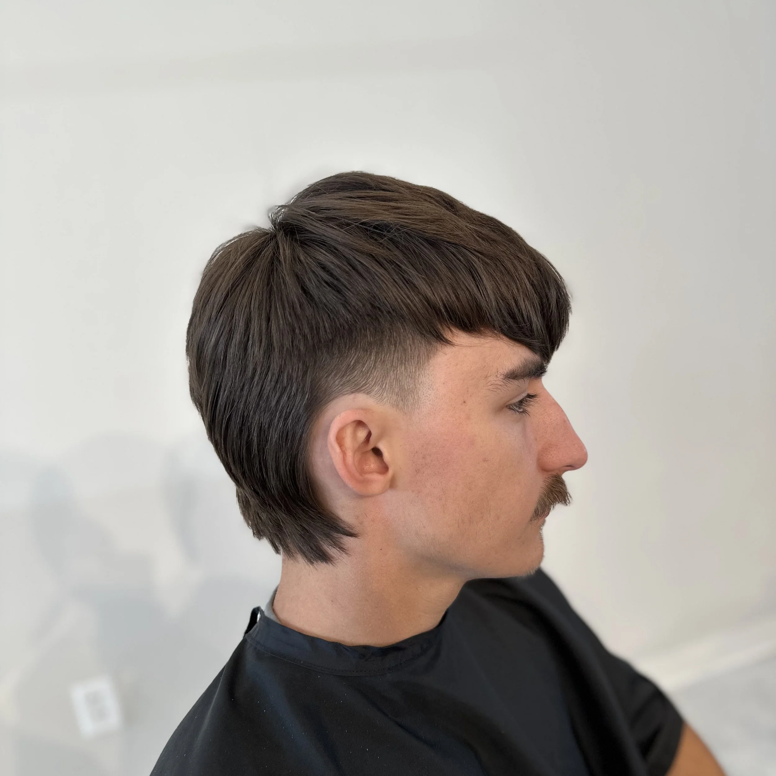 Side profile of a young man with dark brown, layered haircut and short moustache, sitting against a plain white wall.