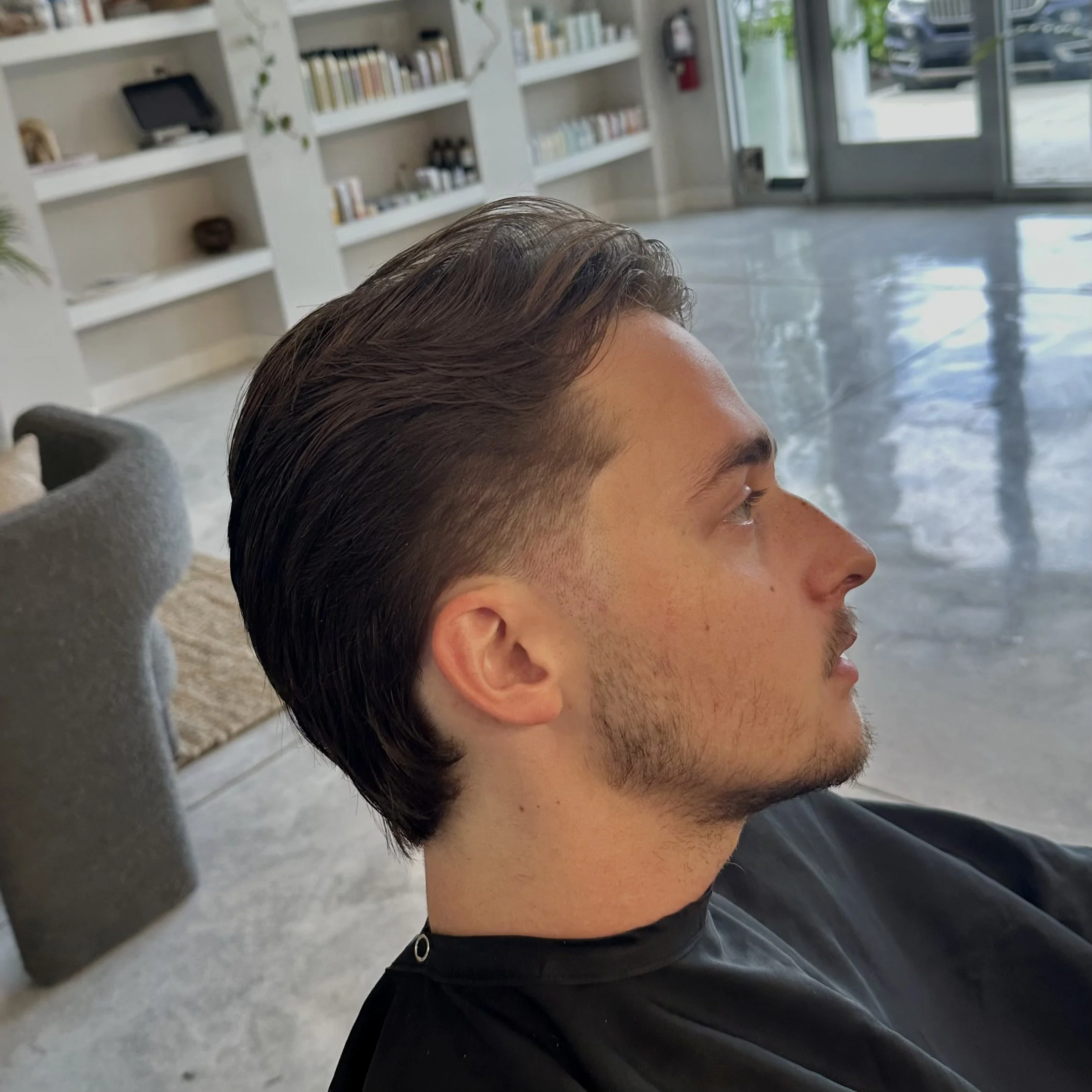 Side profile of a young man with styled brown hair, a mustache, and light beard, sitting indoors in a salon or similar setting with white shelves filled with books or products in the background.