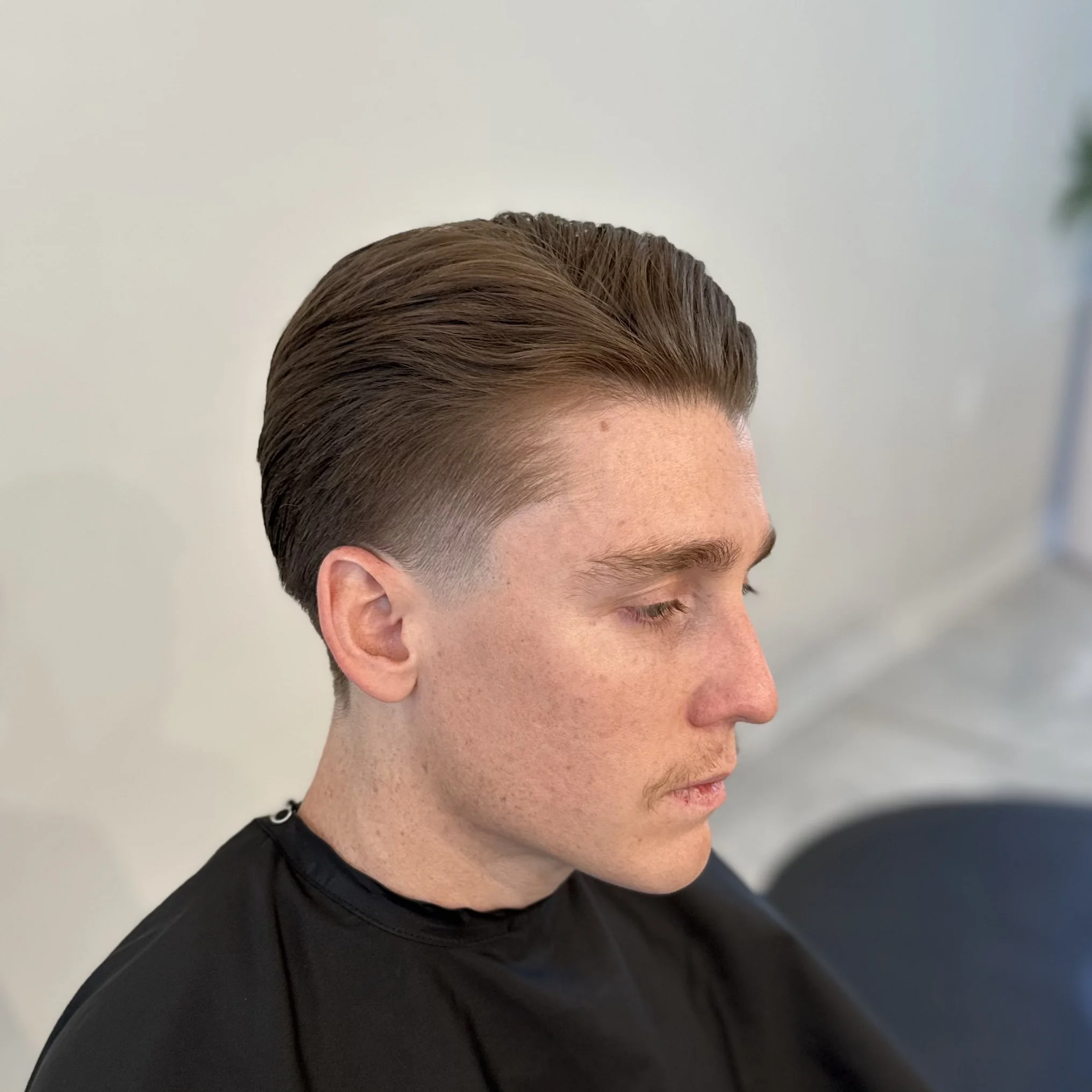 Side view of a young man with a classic slicked-back hairstyle, seated with eyes closed, wearing a black cape in a salon.