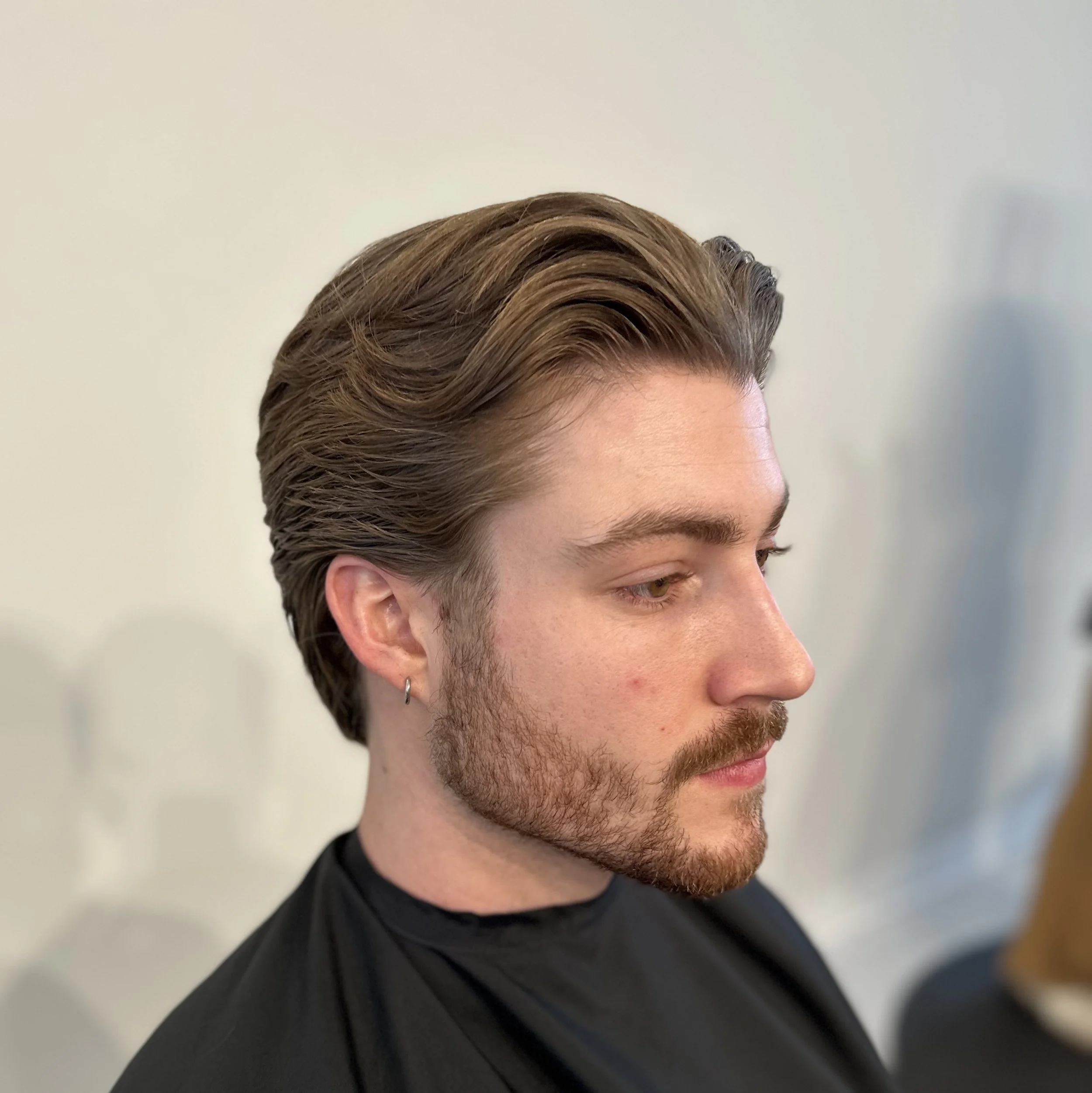 Profile of a young man with styled brown hair, beard, and hoop earring, wearing a black cape, in a salon or barbershop setting.