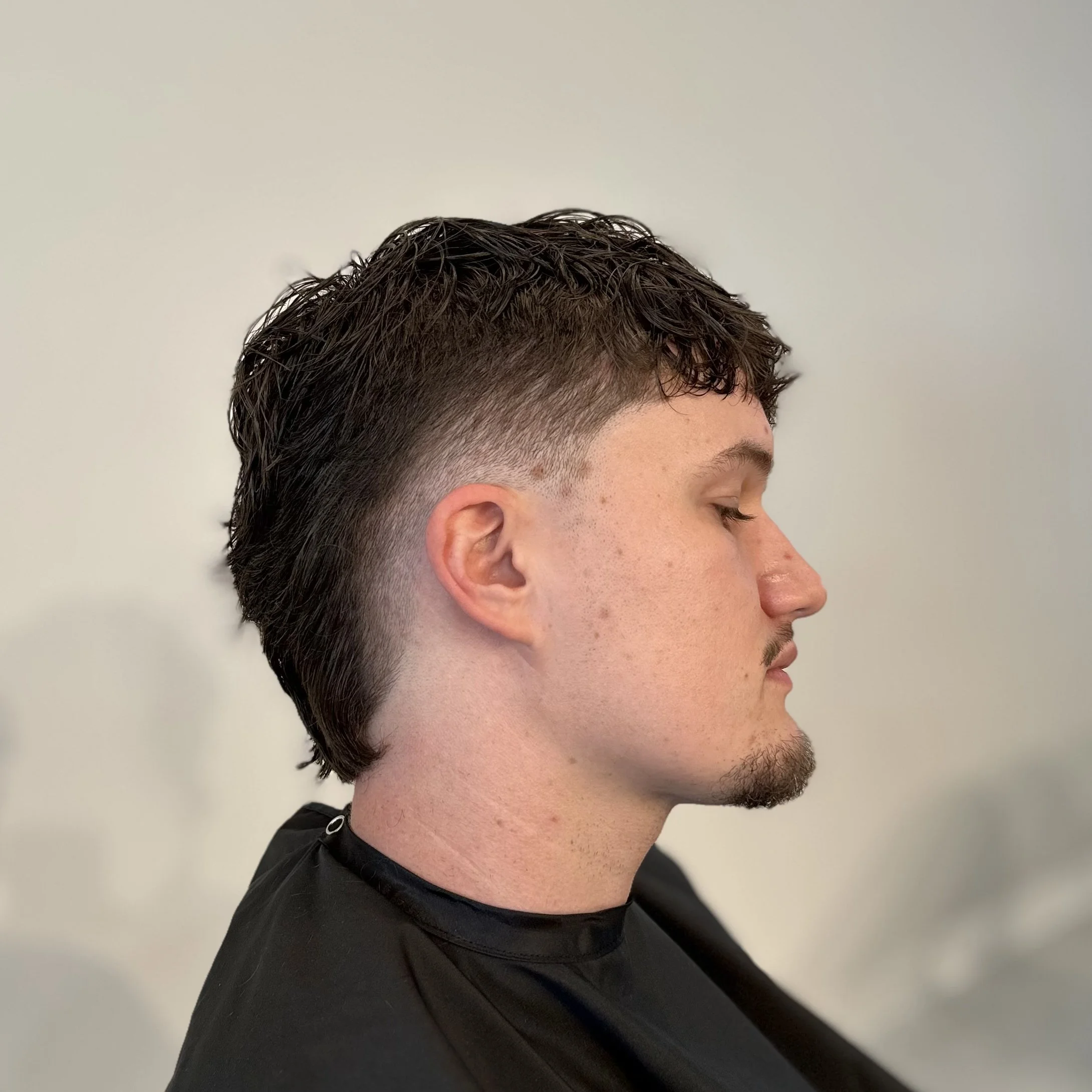 Side profile of a young man with short, wavy dark hair, fading into a lighter undercut, with a trimmed beard and mustache, sitting with eyes closed against a plain, light-colored background.