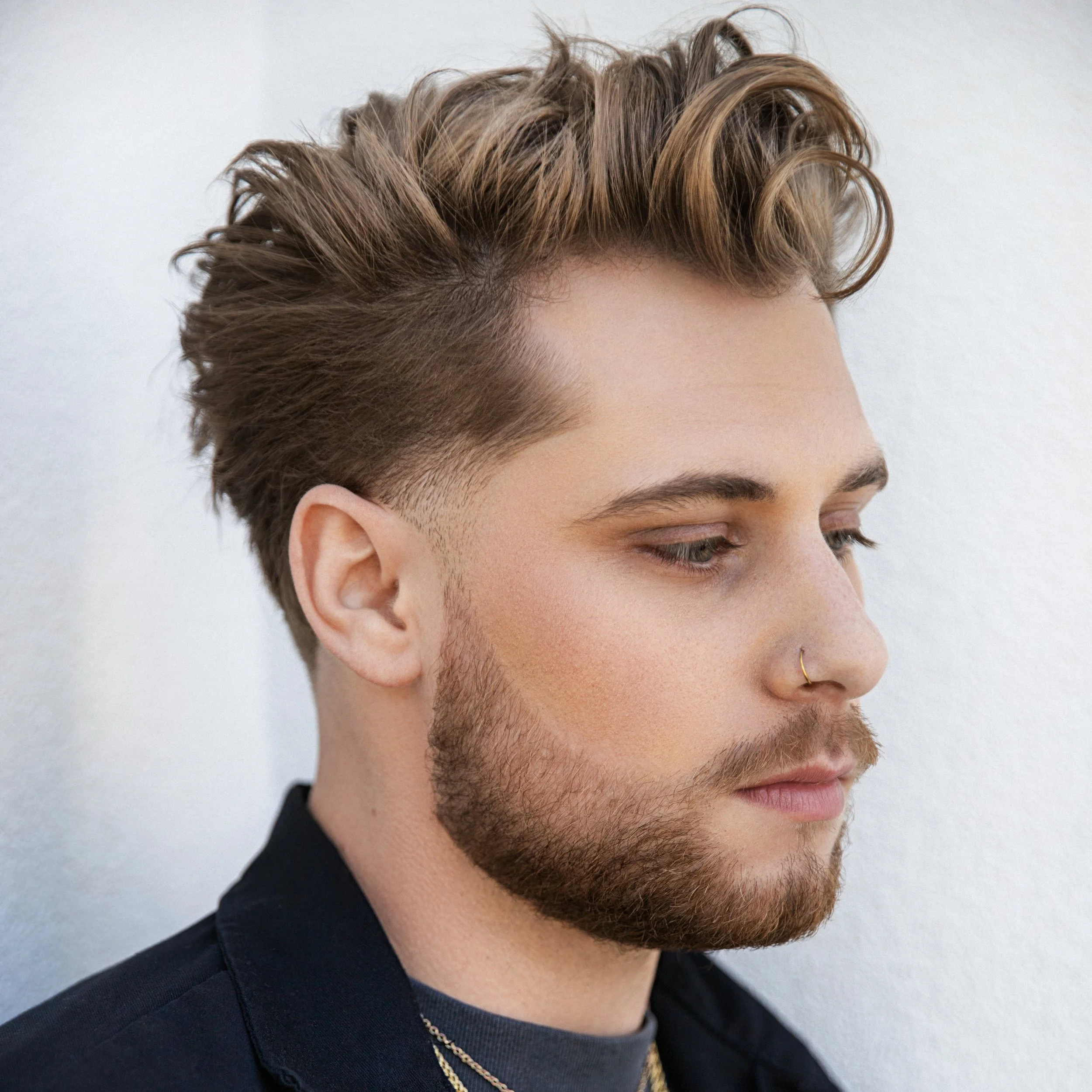 Close-up of a young man with styled brown hair, beard, and a nose ring, looking down, wearing a black shirt and gold chains.