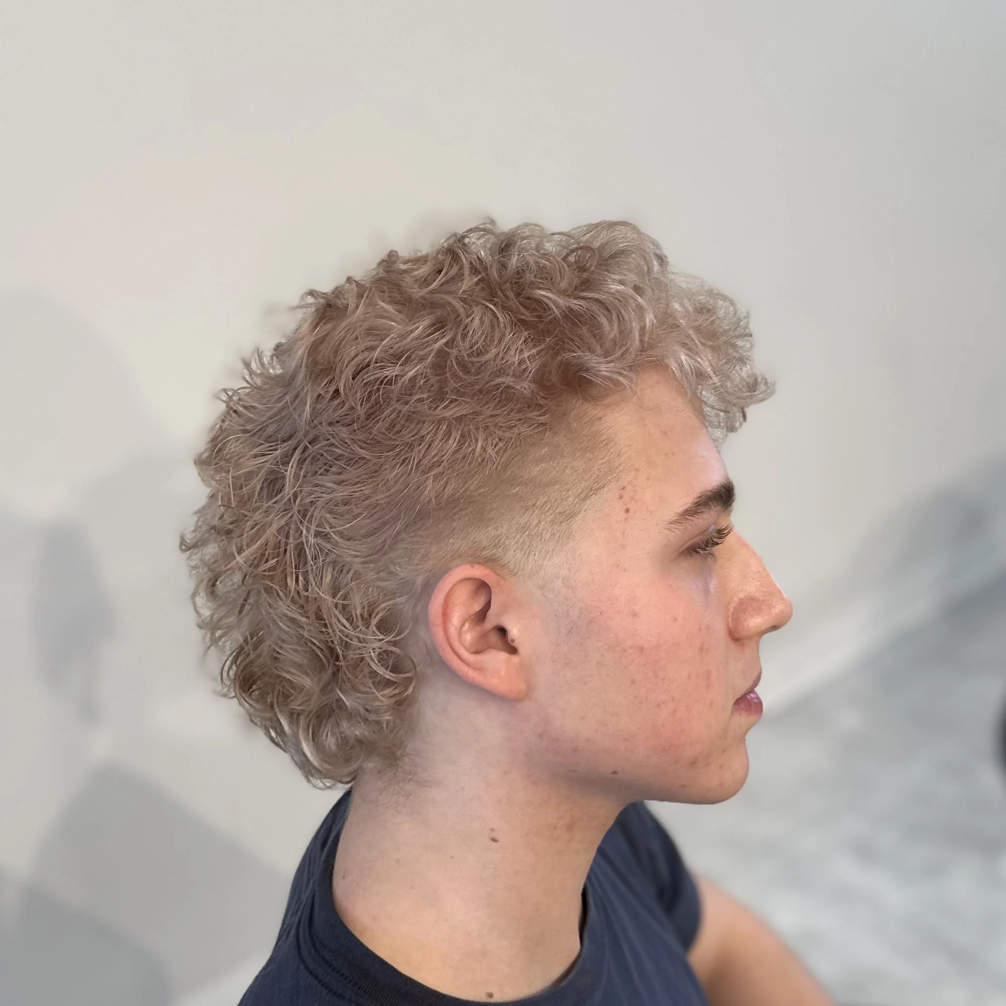 Side profile of a young person with curly, bleached hair and a faded undercut, wearing a dark shirt.
