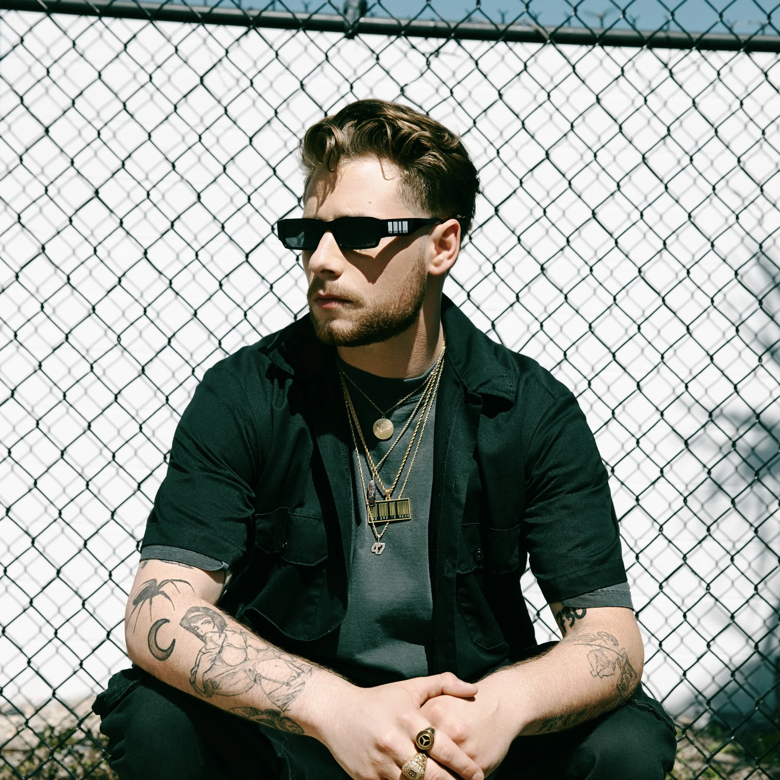 A young man with tattoos on his arms and styled hair, wearing black sunglasses, layered necklaces, a black short-sleeve shirt over a gray T-shirt, sitting in front of a chain-link fence.