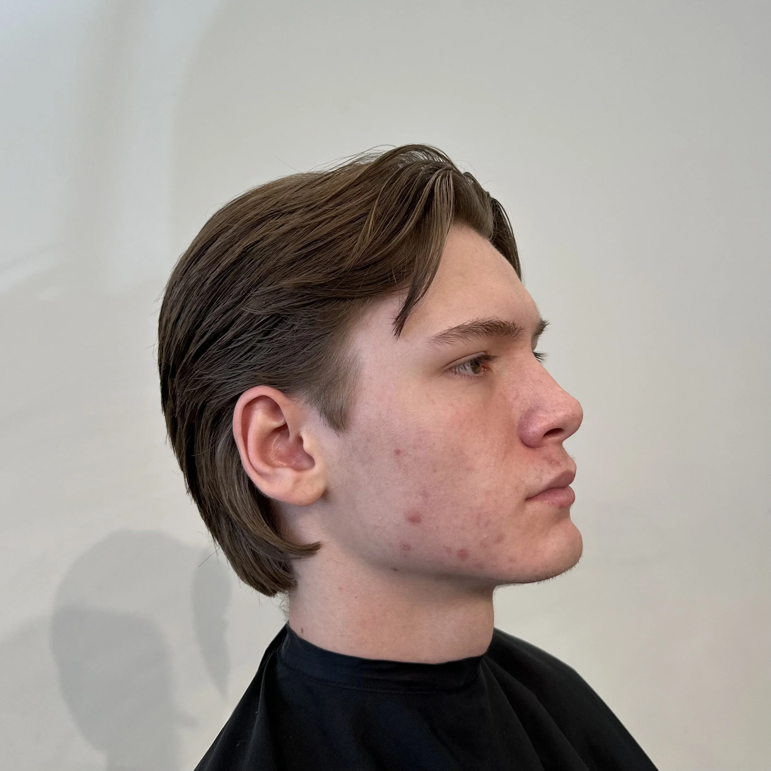 Profile of a young man with straight brown hair, clear skin, and slight acne, facing to the right, against a plain light-colored wall.