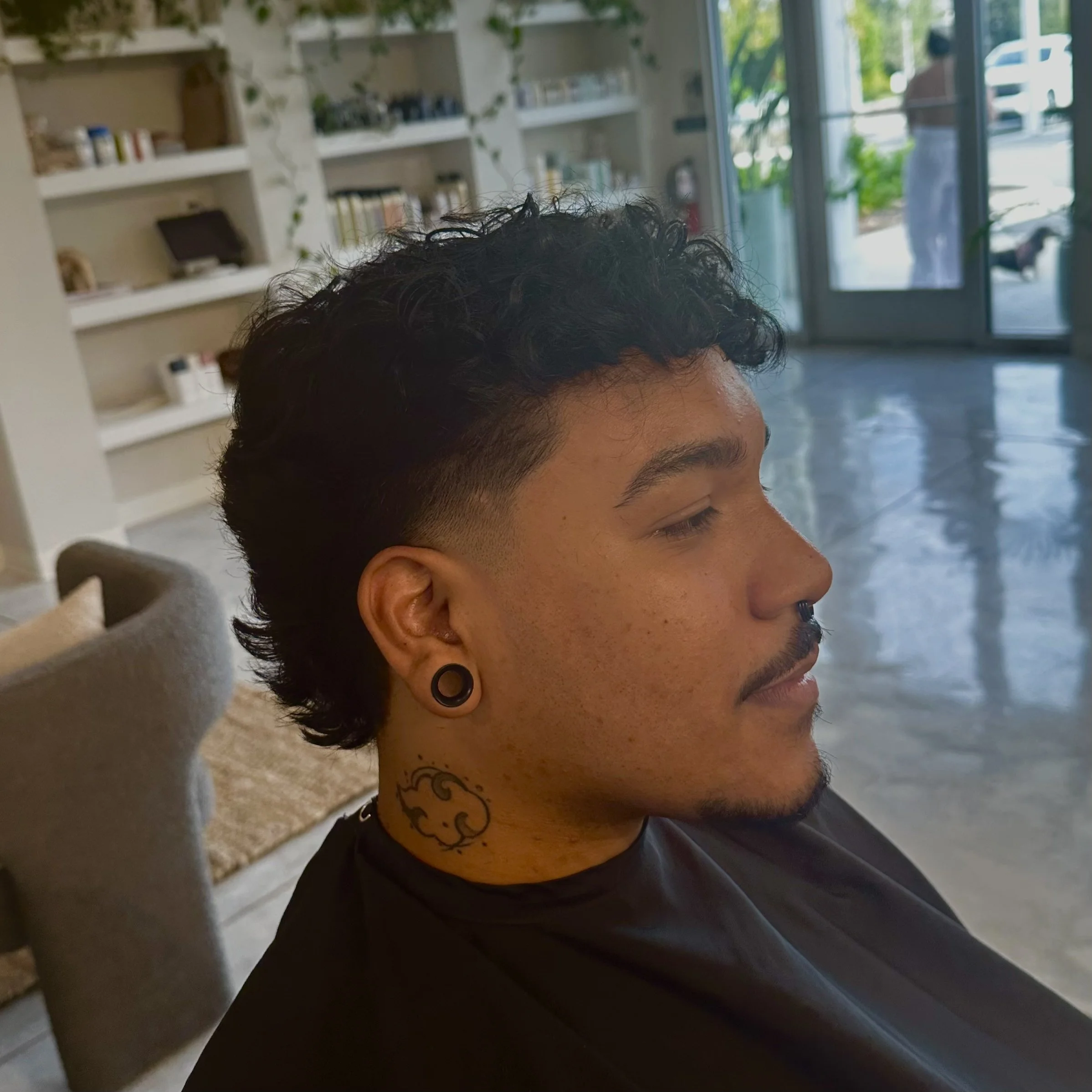 Side profile of a young man with a curly undercut hairstyle, black ear gauges, and a cloud tattoo on his neck, sitting in a salon or barbershop.