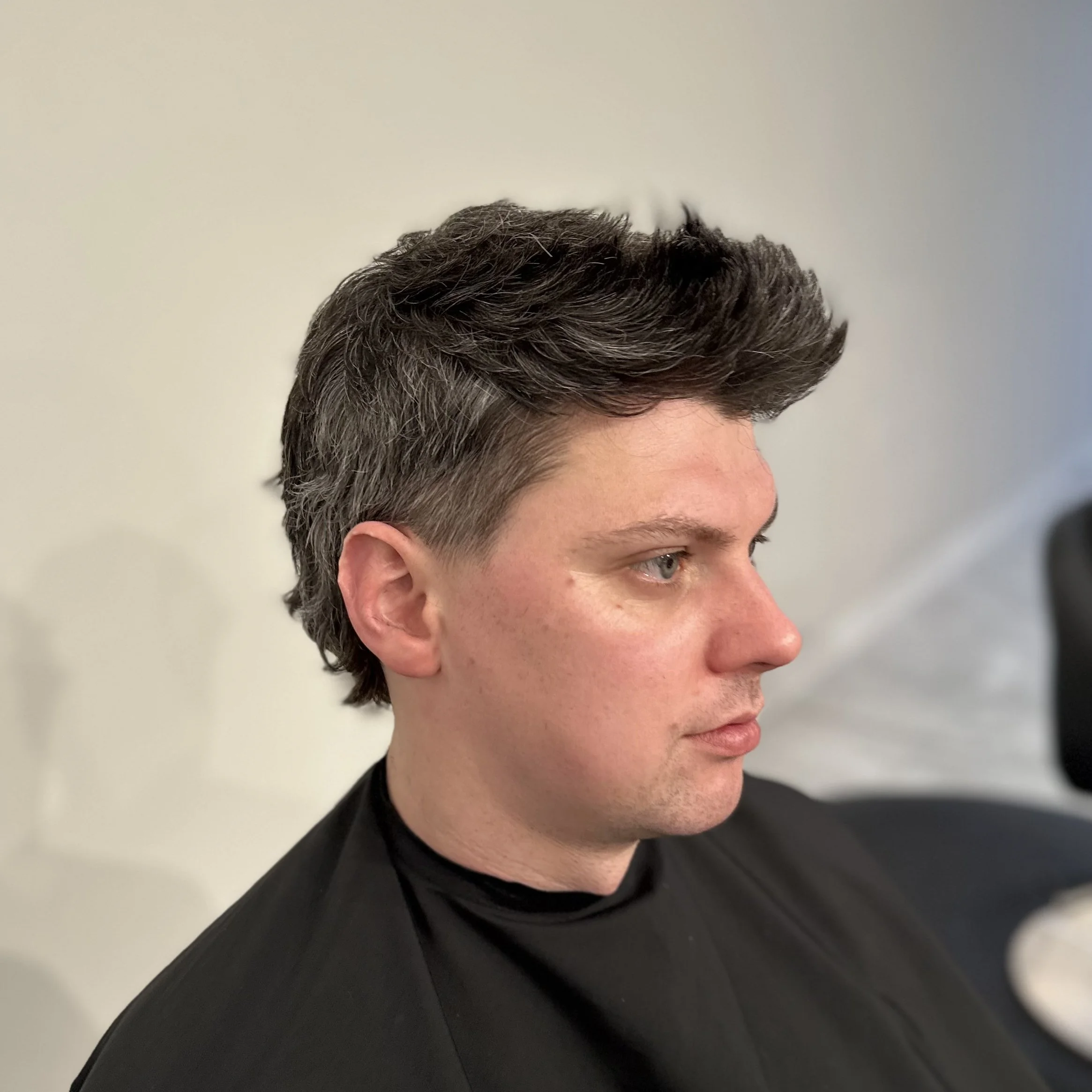 Side profile of a young man with dark, styled hair, wearing a black shirt, sitting against a plain background.