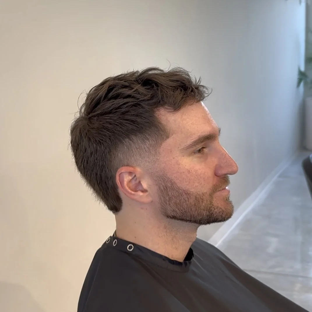 Side profile of a man with a fresh haircut, featuring a fade on the sides and longer textured hair on top, sitting in a salon chair.