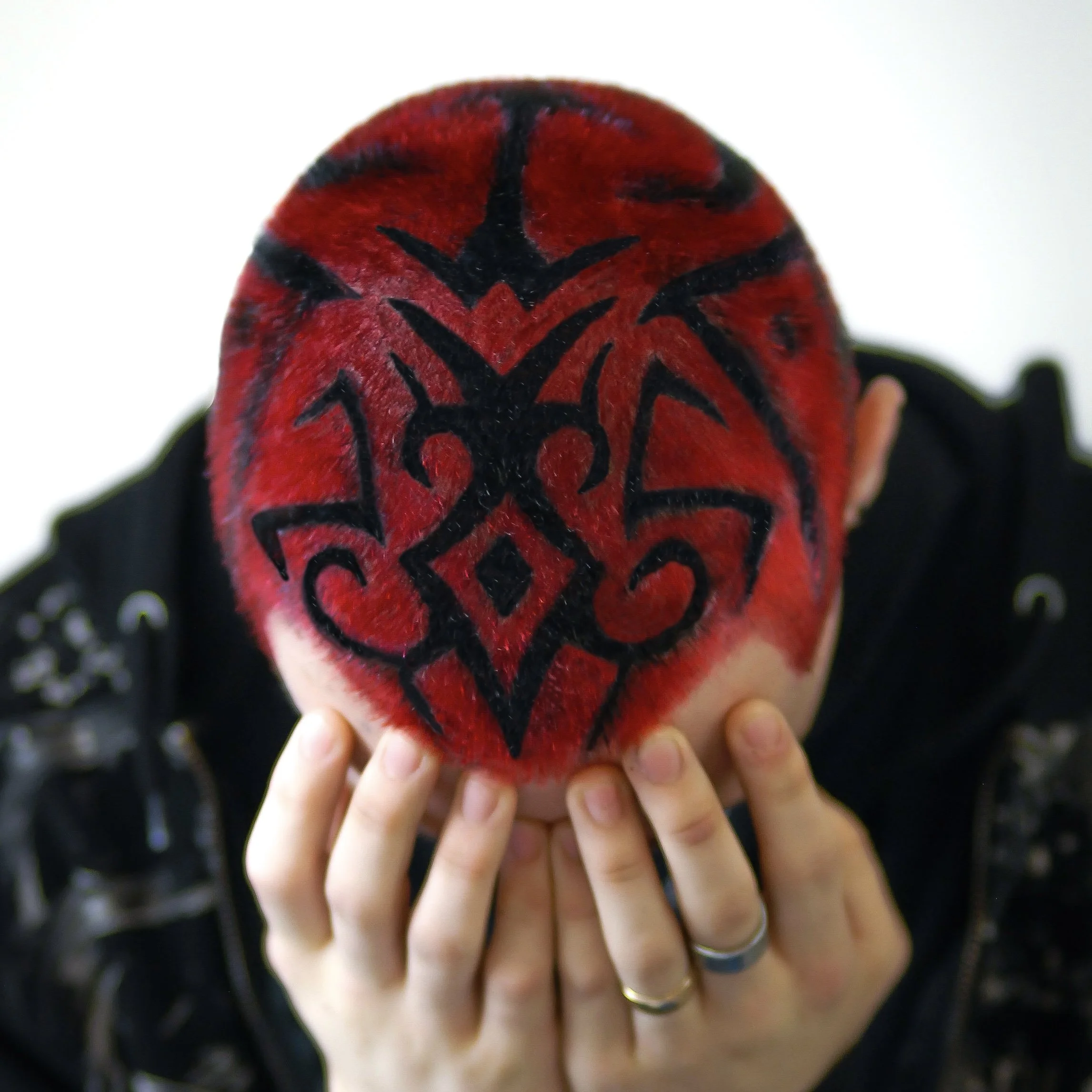 Person with a shaved head featuring a black tribal tattoo design on red hair, holding their face with hands, wearing rings.
