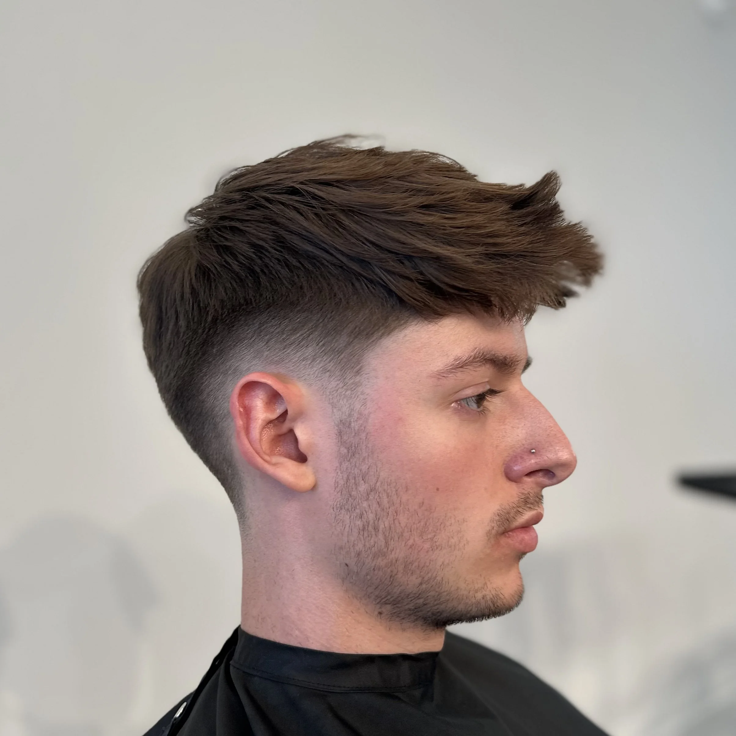 Side profile of a young man with a modern undercut hairstyle, brown wavy hair on top, and a nose piercing, sitting in a salon.
