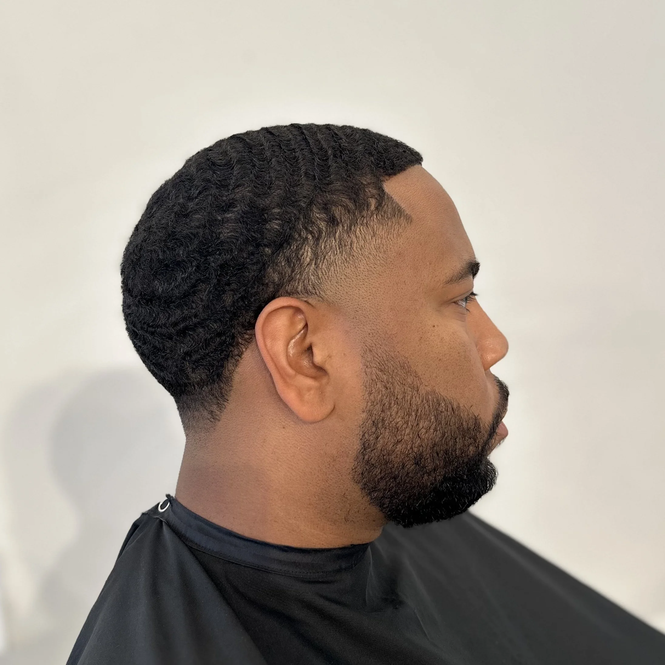 Side profile of a man with a tapered haircut and a beard, wearing a black barber cape.