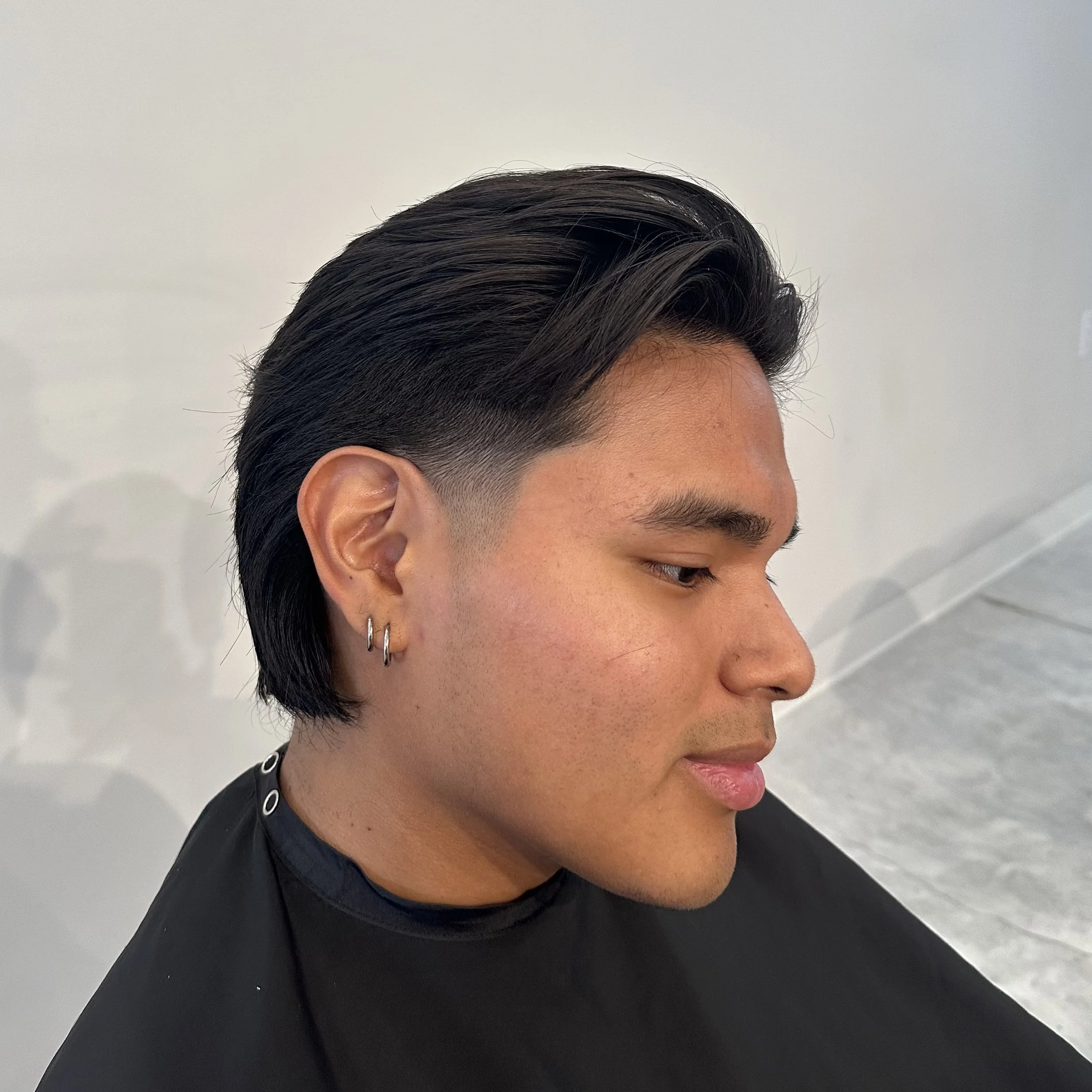 Side profile of a young man with dark slicked-back hair, wearing multiple earrings, and a black cape in a salon.