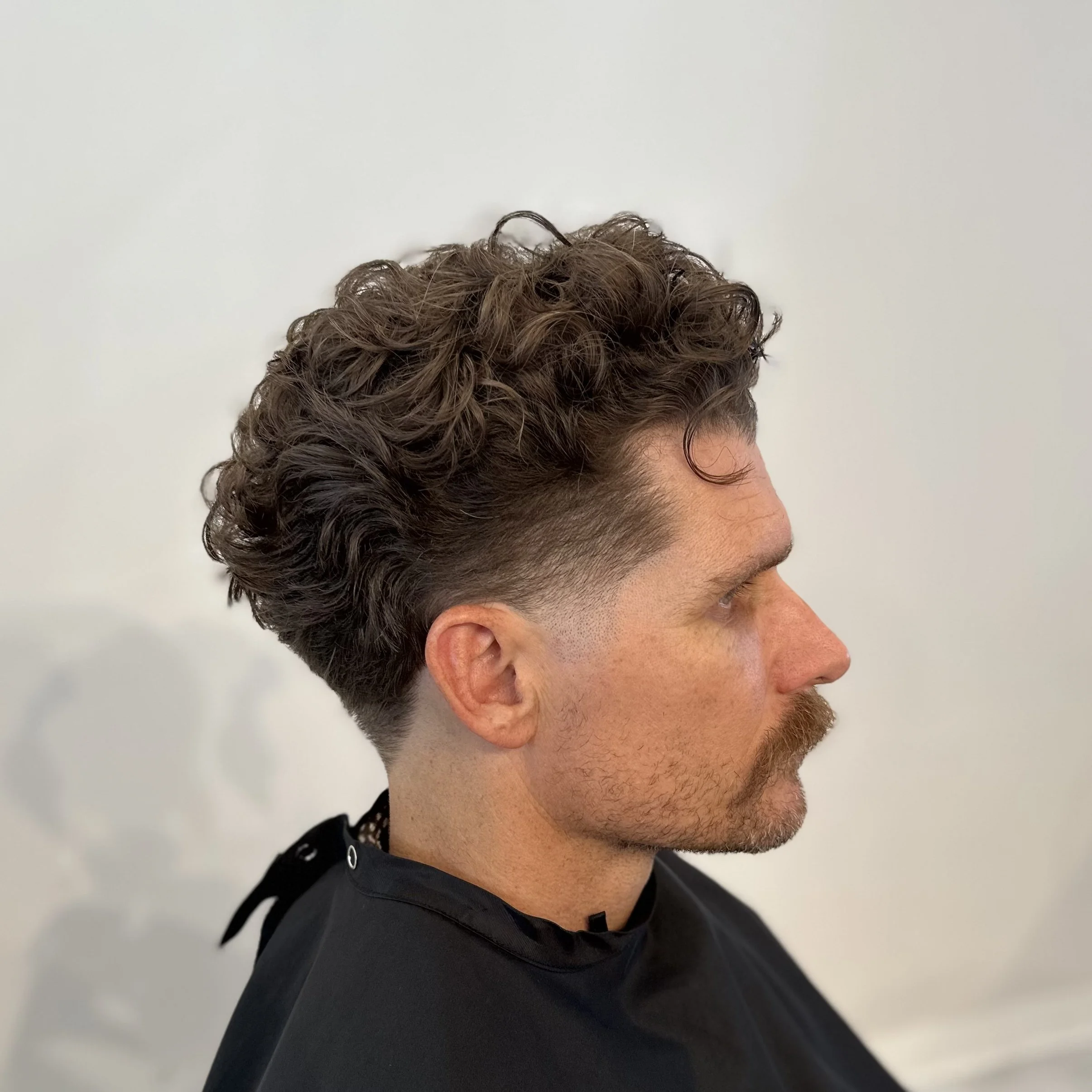 Side profile of a man with a high fade haircut and curly hair on top, wearing a black salon cape