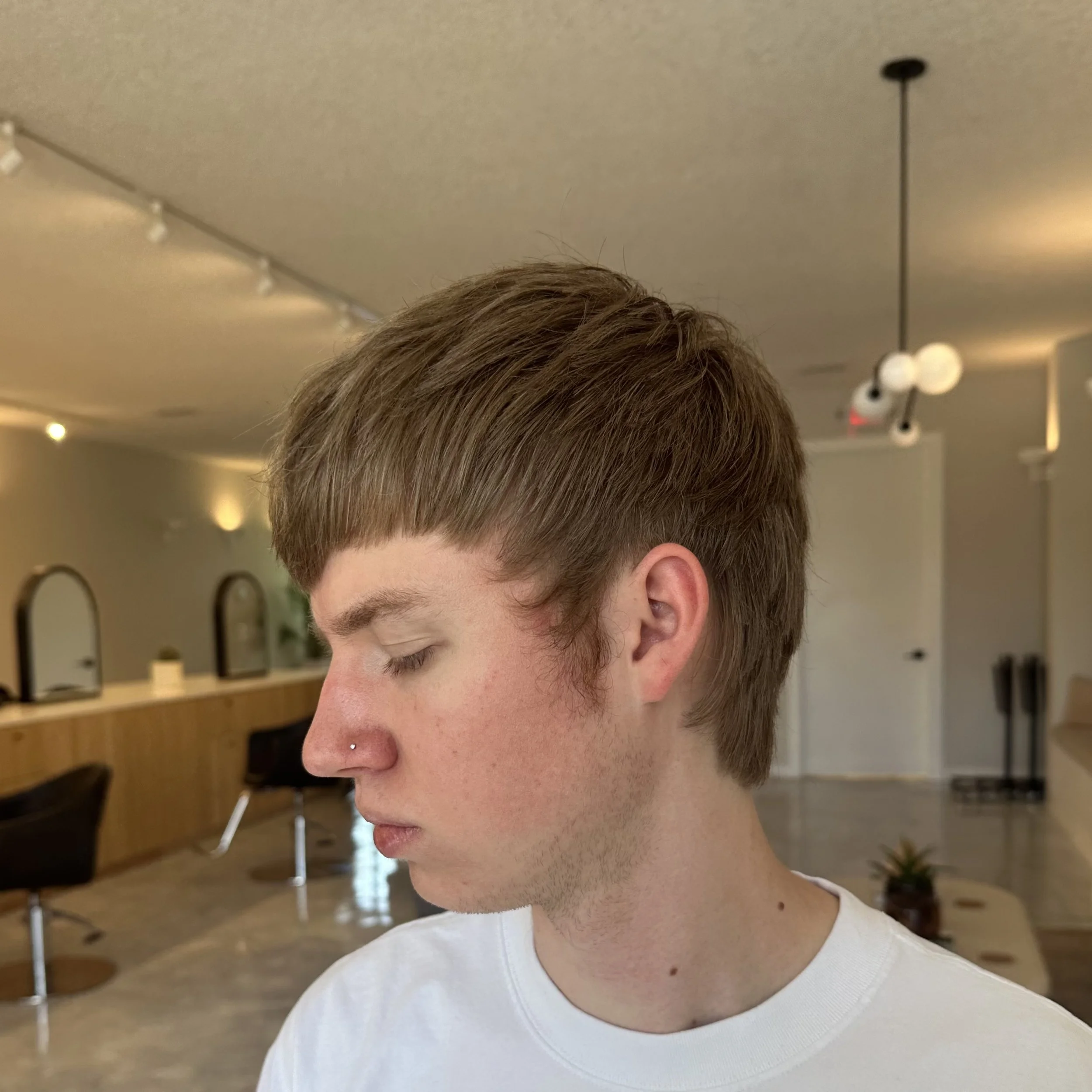 Young man with light brown hair and a nose piercing in an indoor setting, with a modern, minimalistic background including chairs, mirrors, and a light fixture.