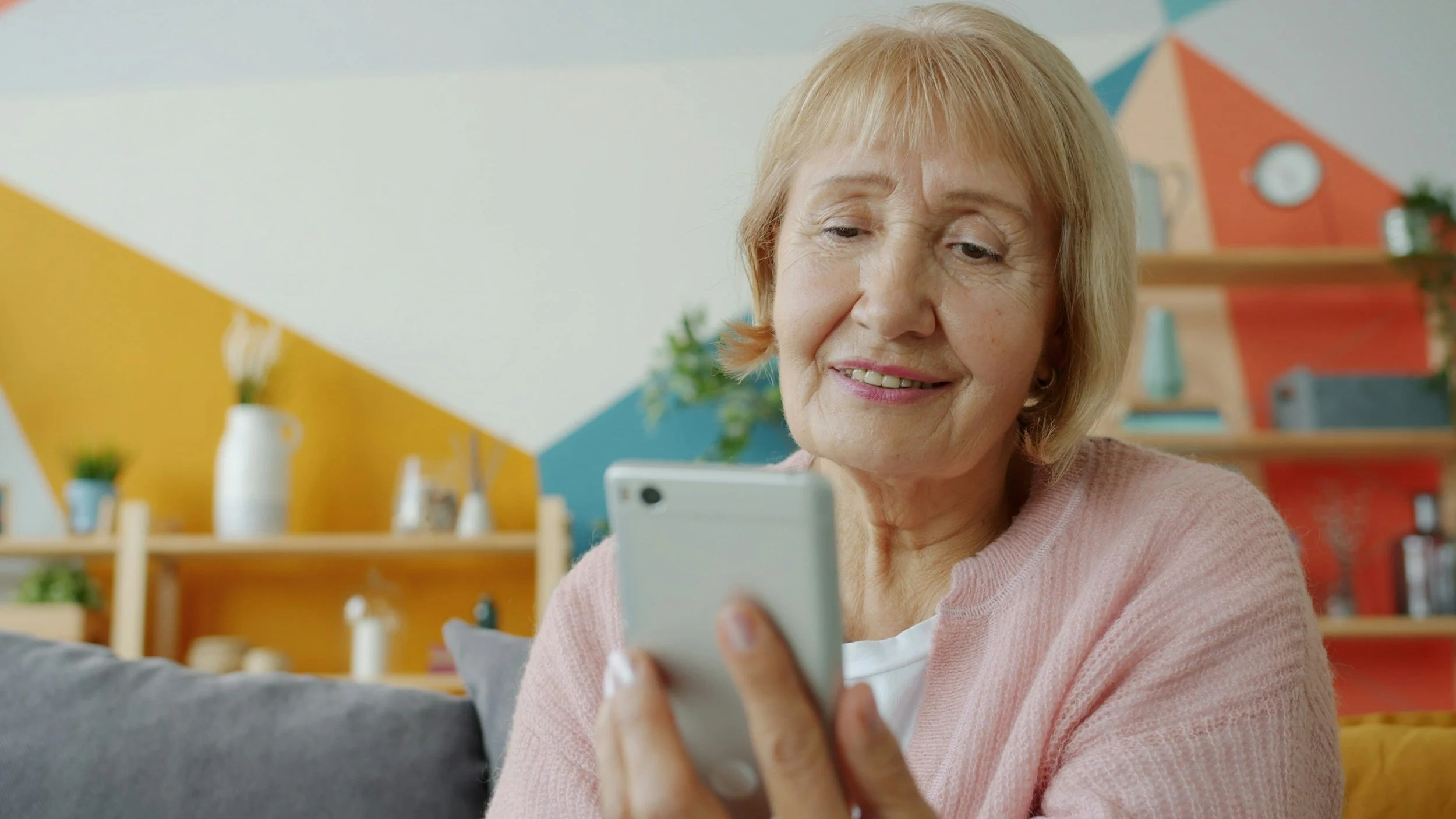 An older woman with blonde hair smiling while looking at her smartphone, sitting on a couch in a colorful room with geometric wall art and decorative shelves.