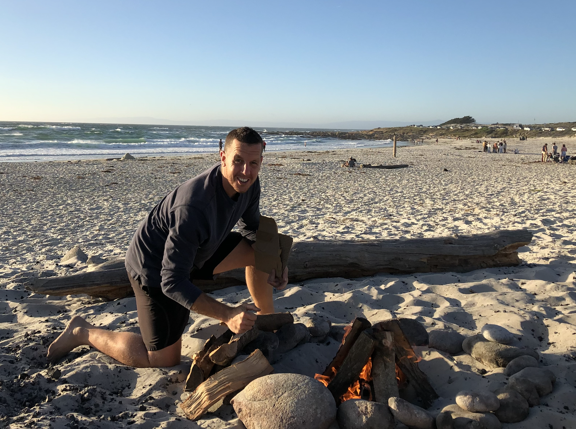 Man kneeling on the beach next to a small fire with logs, smiling and holding a piece of wood, with the ocean and other beachgoers in the background during sunset.