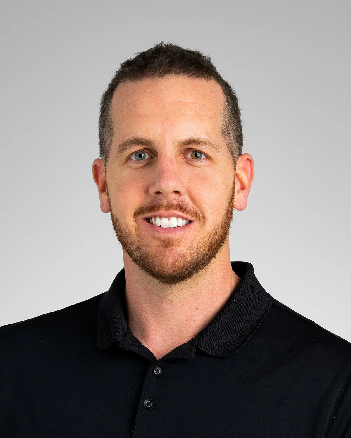A man with short brown hair, a beard, and blue eyes smiling while wearing a black collared shirt. The background is plain and gray.