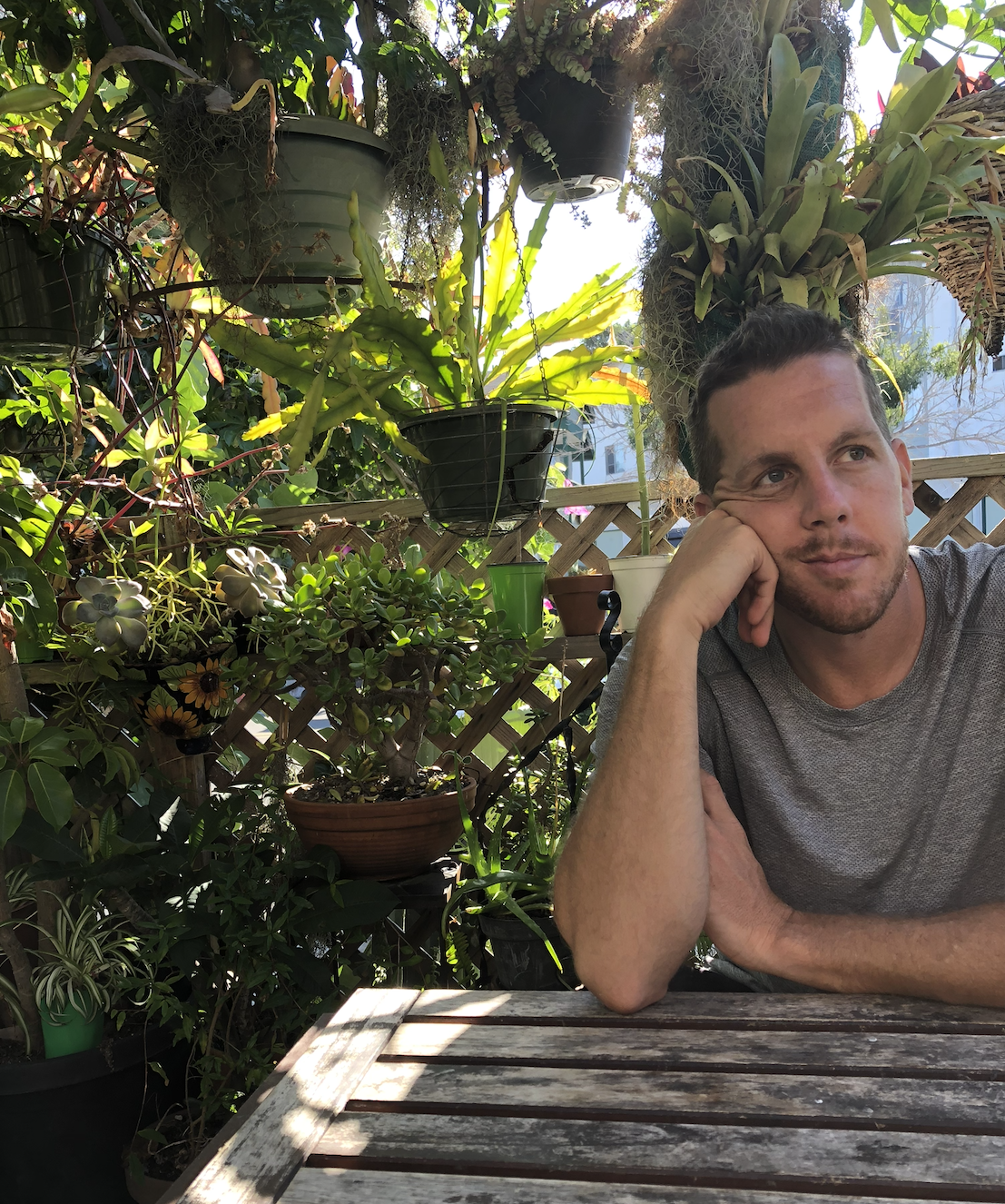 A man sitting at a weathered wooden table in a lush garden or patio area with various potted plants, hanging baskets, and greenery.
