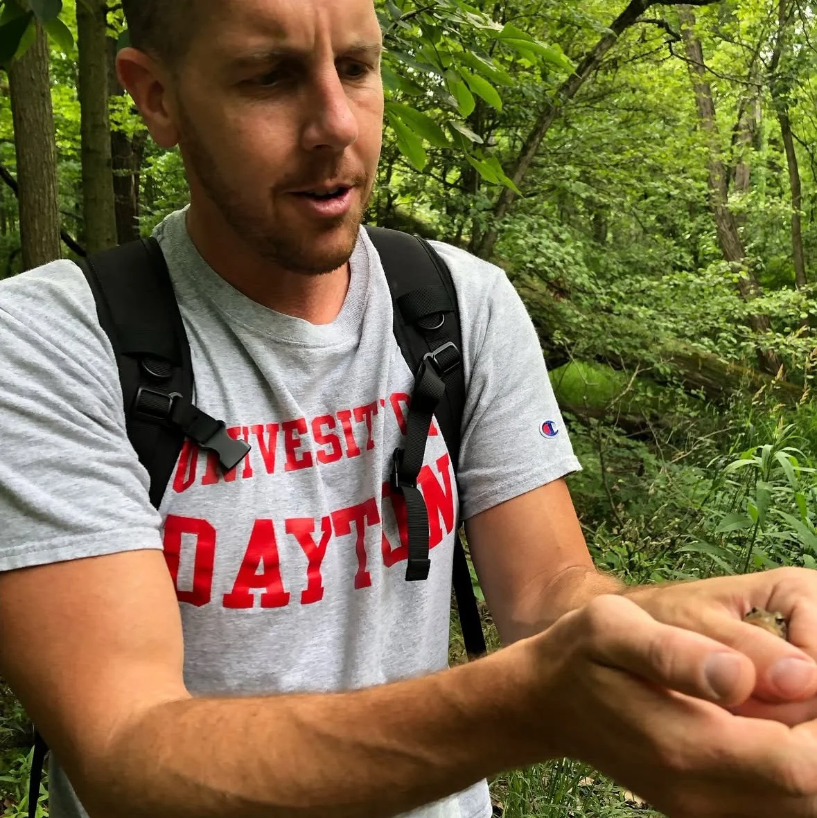 Man in a gray T-shirt with red text and a black backpack in a forest, holding a small animal or creature with both hands.