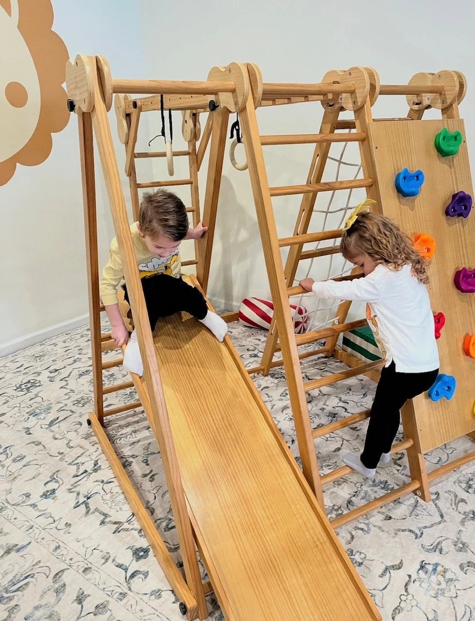 Two children playing on indoor wooden climbing structures, including a slide, ladder, and climbing wall with colorful holds.