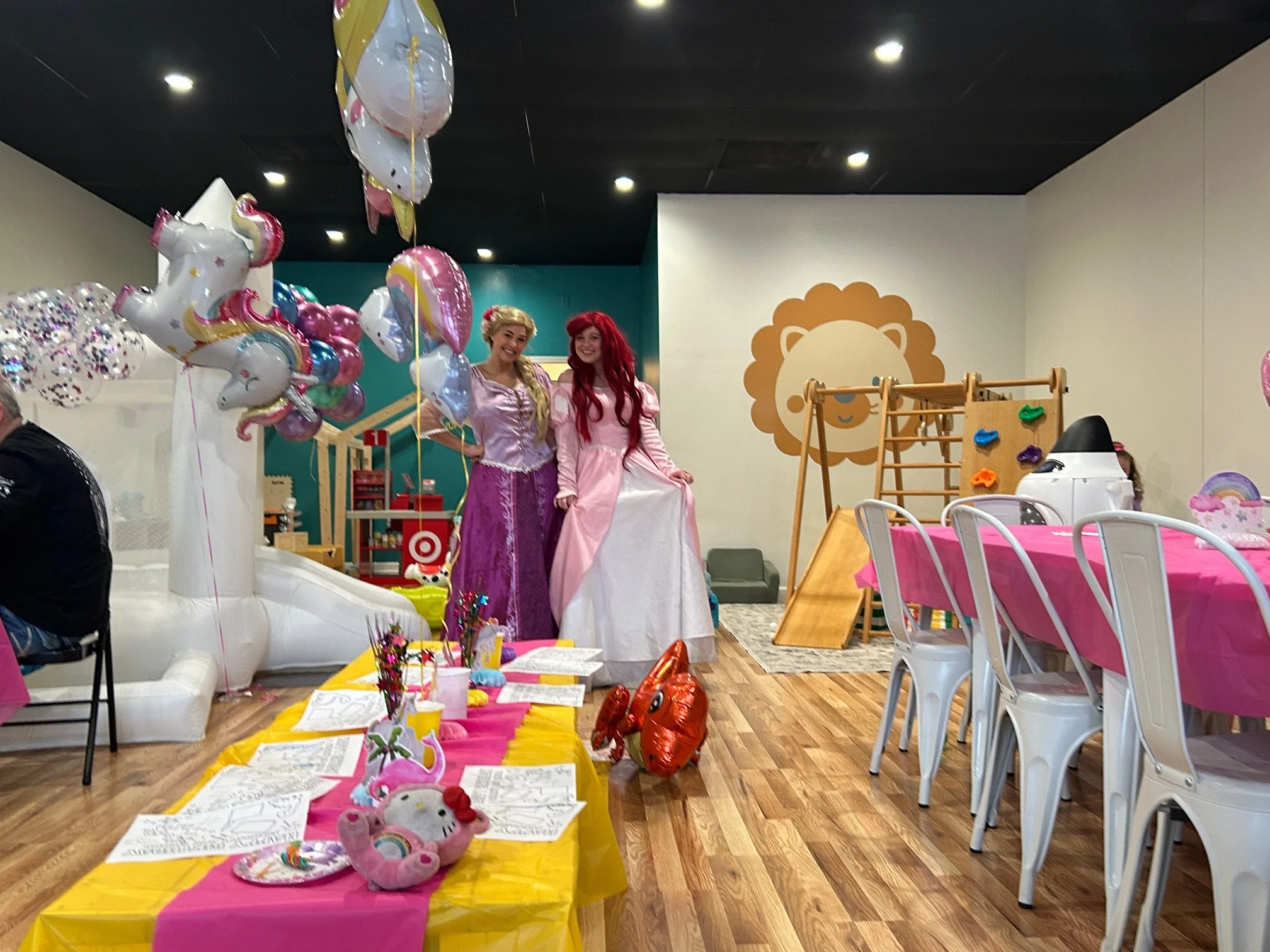 Two women dressed as princesses, one with blonde hair and a pink gown, and the other with red hair and a pink gown, standing in a decorated party room with balloons, tables, and children’s toys.