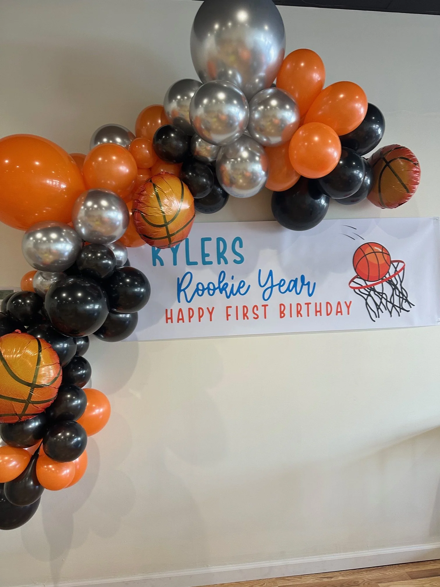 Decorative sports-themed balloon arch with orange, black, silver, and basketball-shaped balloons, and a banner reading "Kyler's Rookie Year Happy First Birthday" with a basketball hoop graphic.