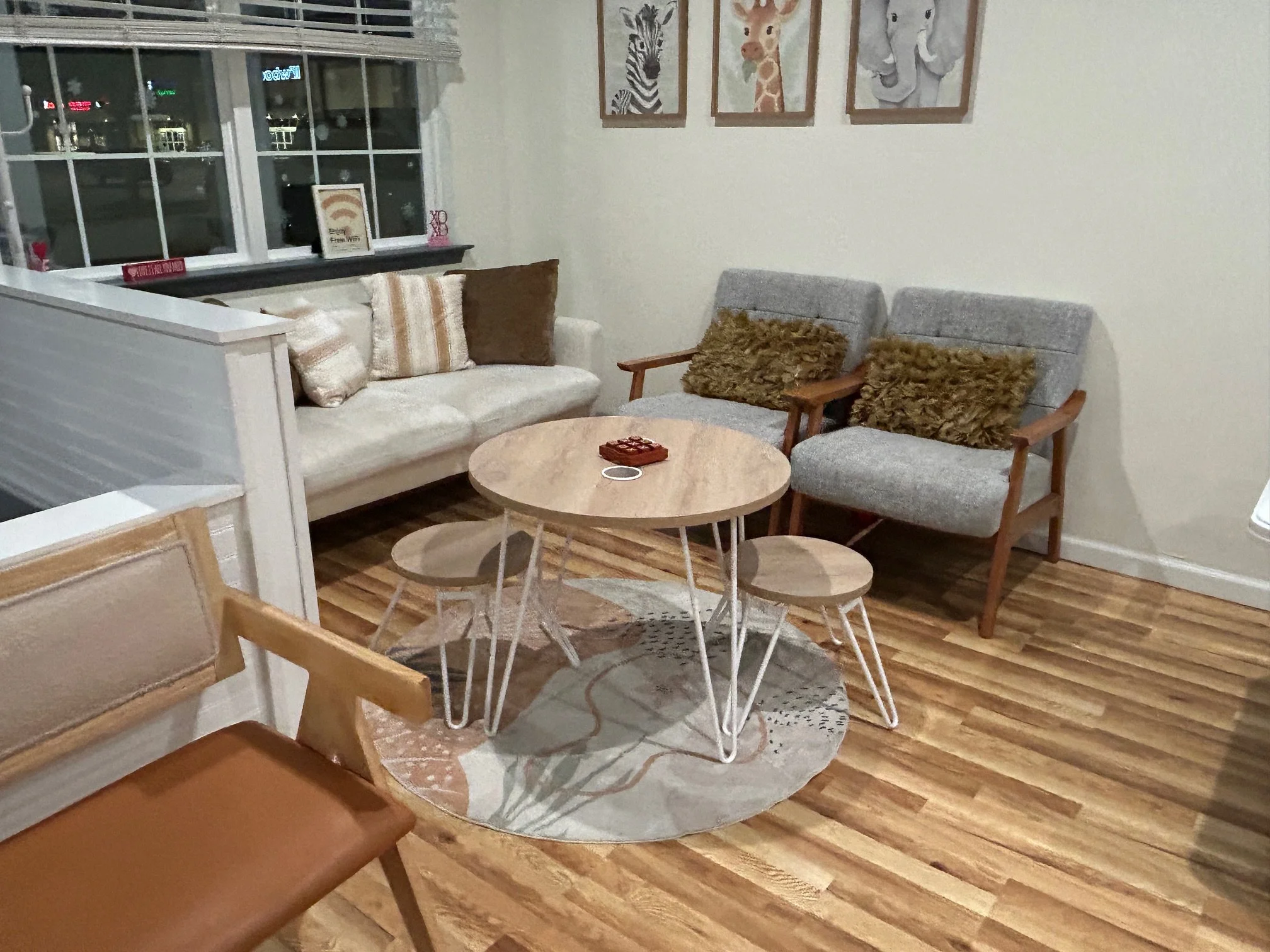 Living room with two armchairs, a sofa, and a round coffee table on a patterned rug. Wall decorations include animal-themed artwork, and the room has wood flooring. A window shows a night scene outside.