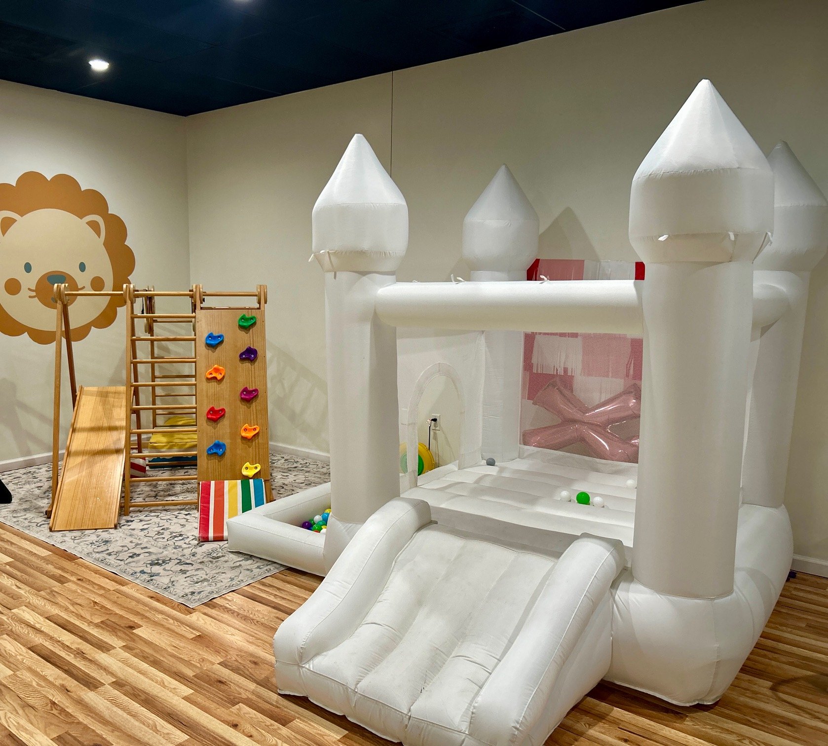 Indoor children’s play area with a white inflatable castle featuring turrets, a slide, and colorful balls inside. To the left, there is a wooden climbing structure with a slide, rope ladder, and colorful climbing holds. On the wall, there's a lion fa