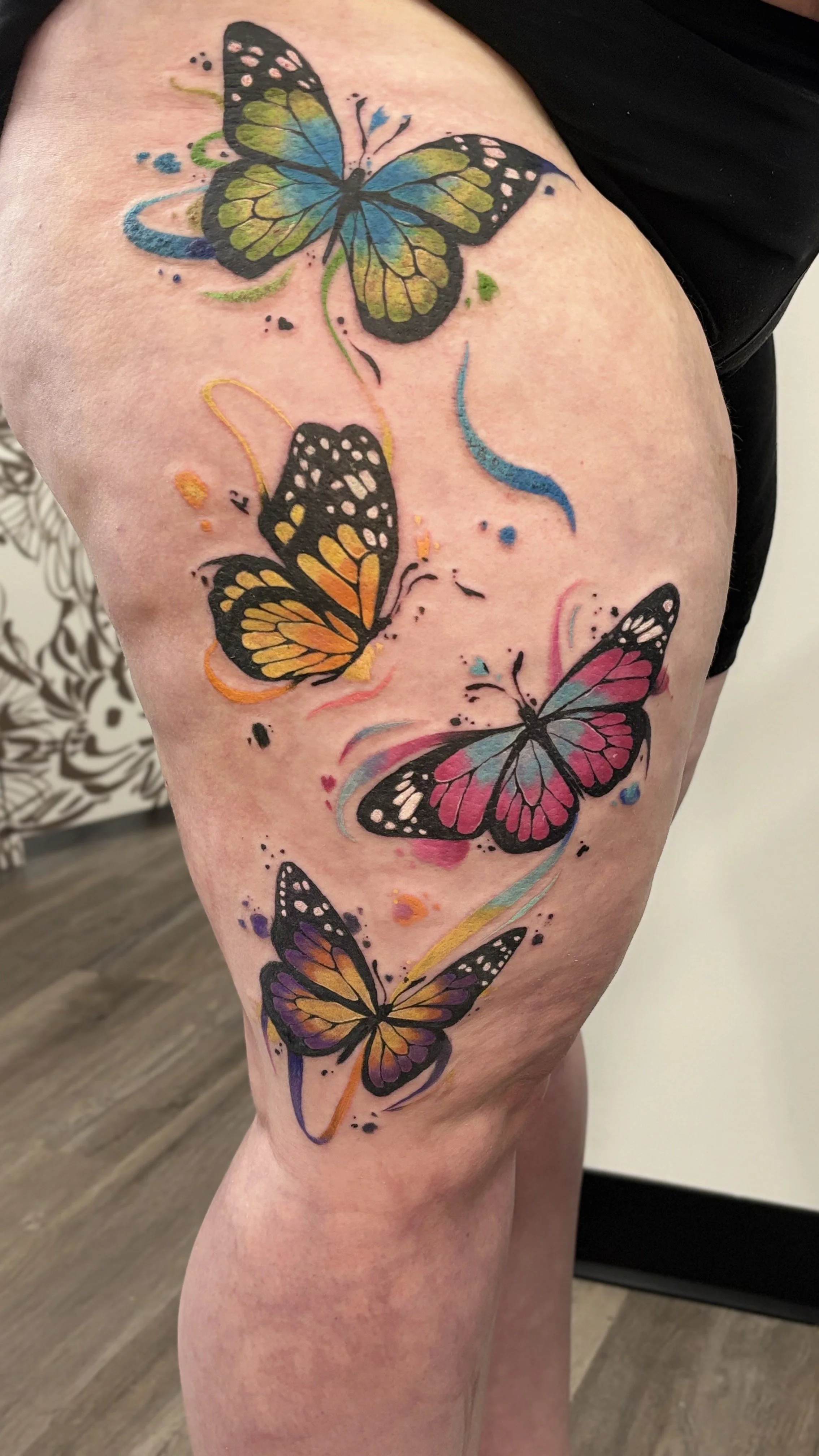 Colorful tattoo of five butterflies with black outlines and vibrant pink, orange, yellow, blue, and purple wings, accompanied by splashes of watercolor-style background on a person's thigh.