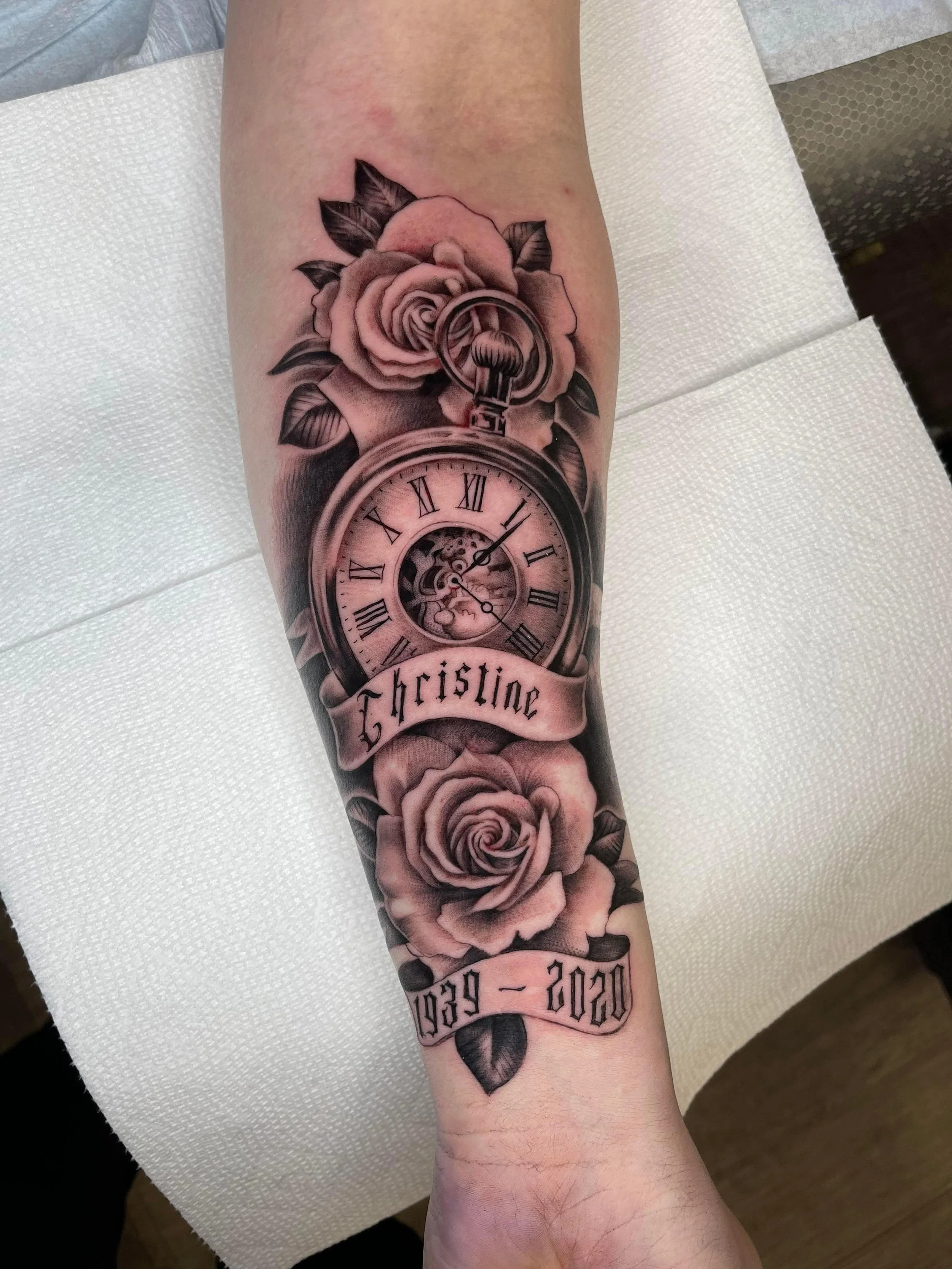 Tattoo featuring a pocket watch, two roses, and a banner with the name 'Christine' on a forearm.
