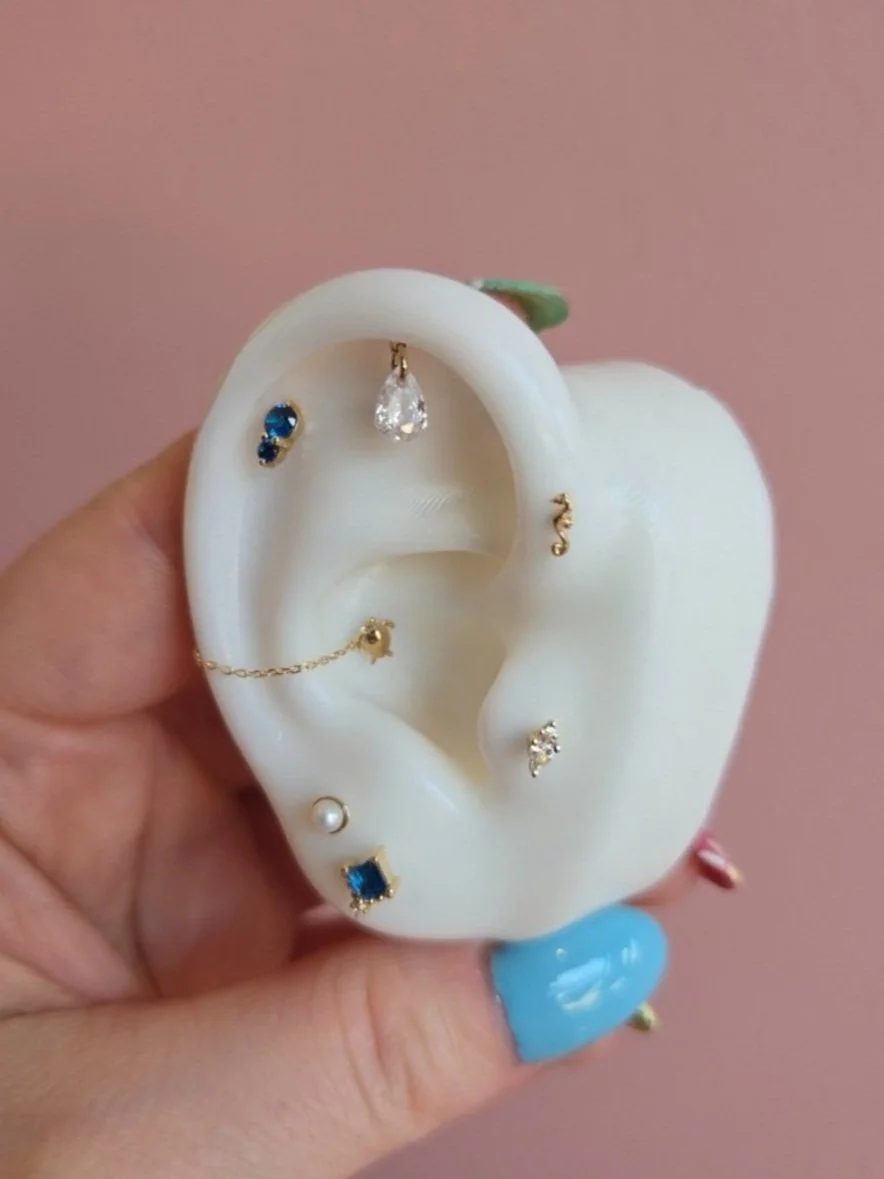 A decorative artificial ear-shaped jewelry holder with multiple earrings and a chain earring displayed on it, held by a hand with painted nails.