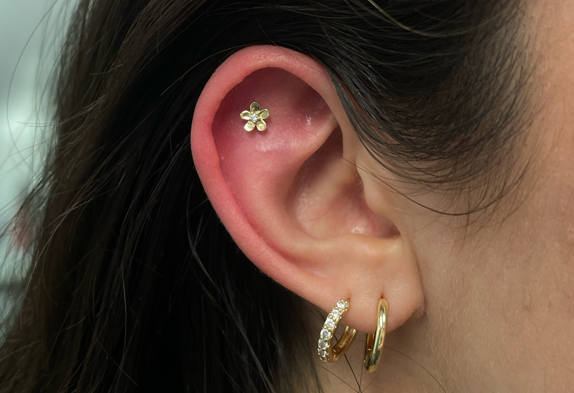 Close-up of a person’s ear with three gold earrings: a small flower-shaped earring with a gemstone, a small hoop with diamonds, and a plain gold hoop. The person has dark hair partially covering the ear.