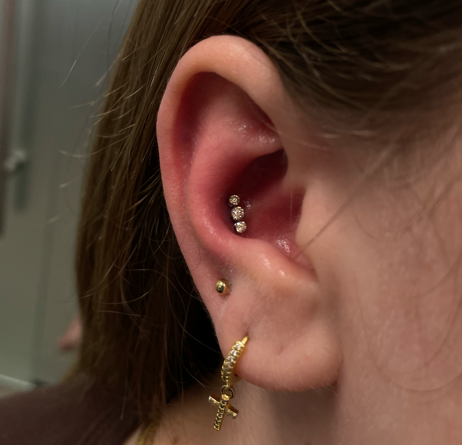Close-up of a person's ear with multiple earrings including three stud earrings with stones, a small gold ball stud, and a gold hoop earring with a cross and the word 'faith'.