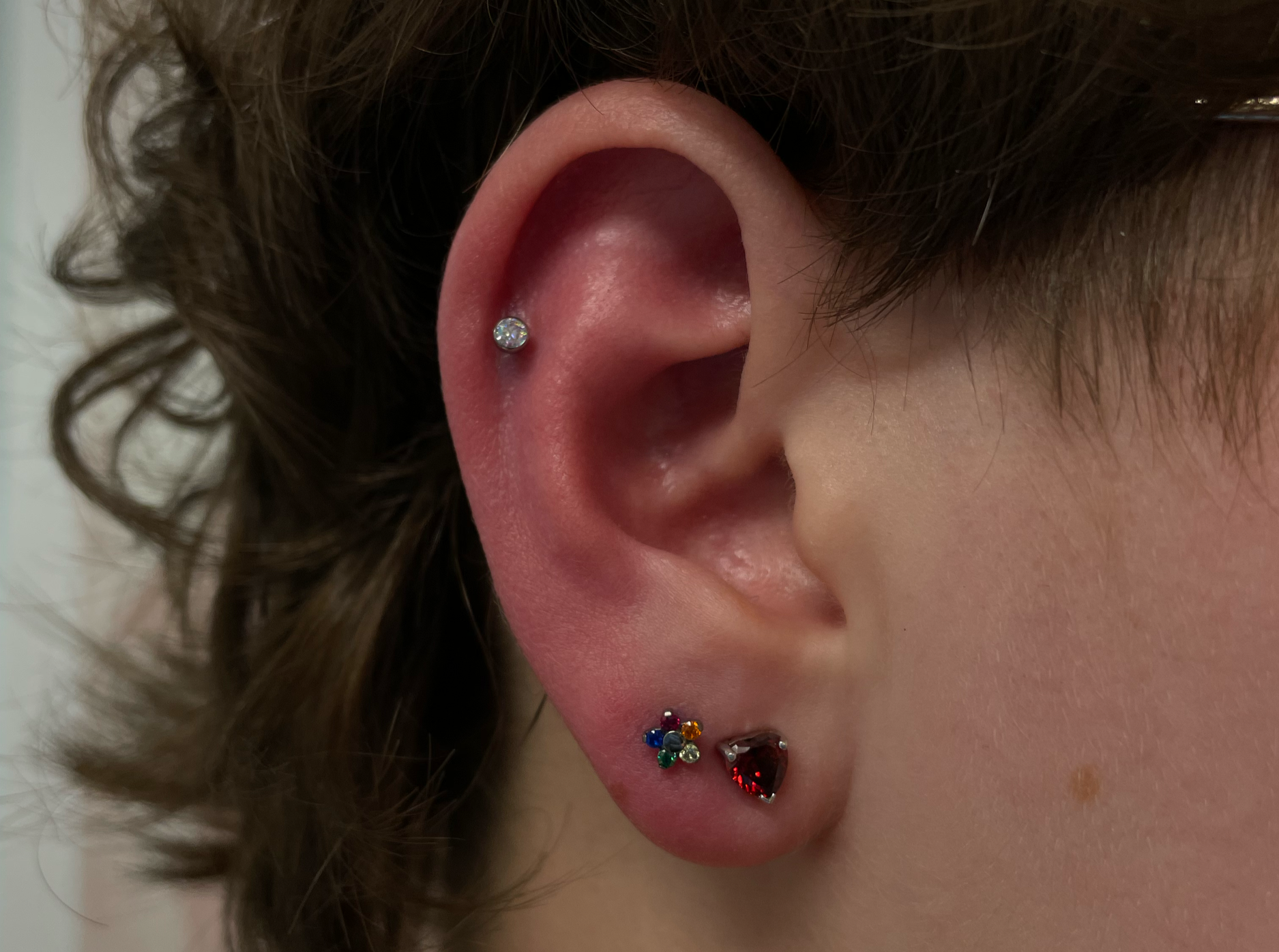 Close-up of a person's ear with four earrings: a small clear crystal in the upper cartilage, a cluster of small multicolored stones in the middle, and a large red gemstone in the lower lobe.