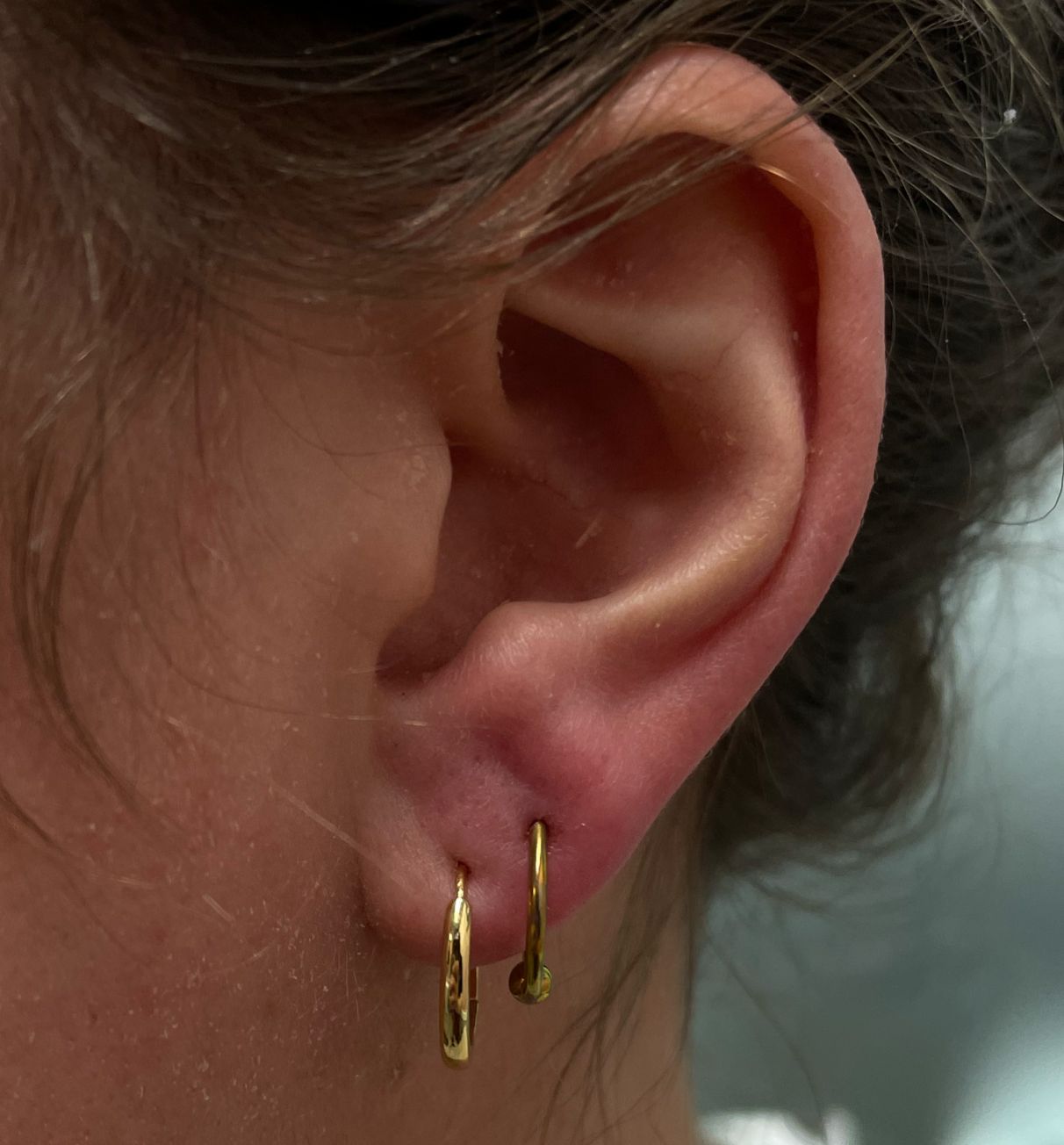 Close-up of a person's earlobe with two small gold hoop earrings, brown hair, and light skin.