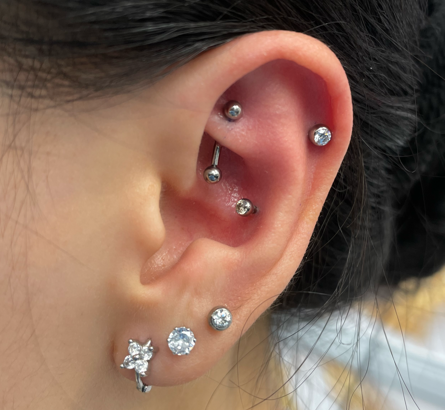 Close-up of a person's left ear with multiple pierced earrings, including a flower-shaped design, a large clear stone, a small gold ball, a stud with a diamond-like gem, and a curved cartilage piercing with a small silver ball.