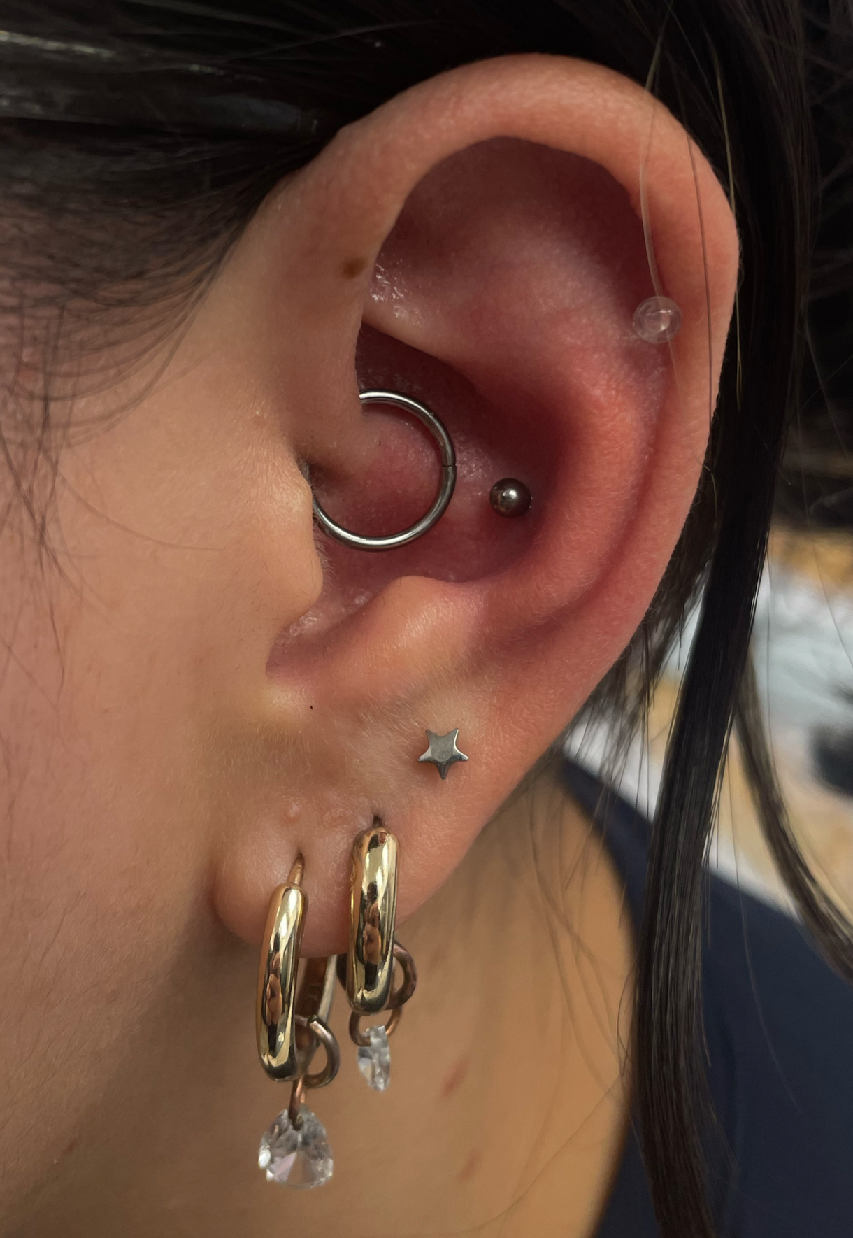 A close-up of a person's ear with multiple piercings, including a large gauge hoop, a small silver ball, a star-shaped stud, and two gold hoops with dangling crystal charms.