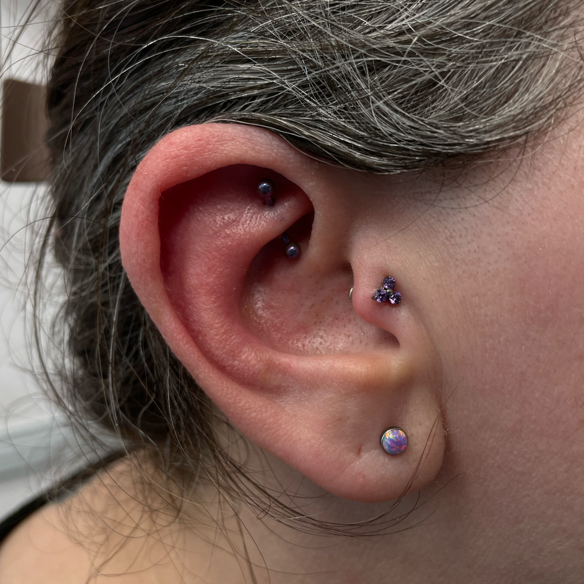 A close-up of a person's ear with multiple piercings, including a helix piercing with a dark ball stud, a rook piercing with a dark ball stud, a nostril piercing with a purple gem stud, and a lobe piercing with an iridescent round stud.